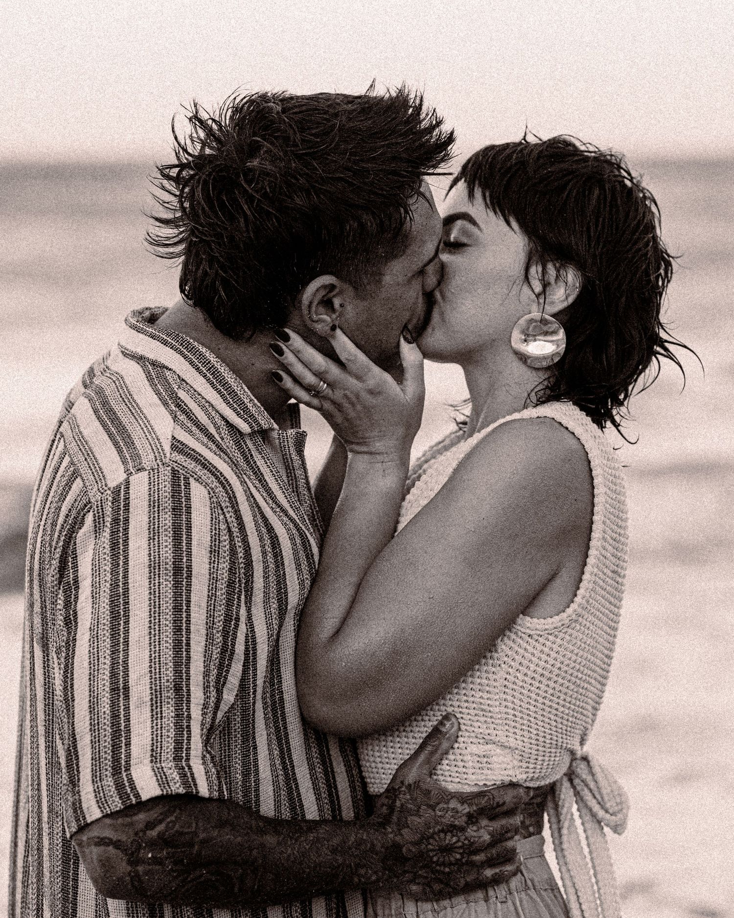 A couple shares a kiss on the beach in a black and white engagement photo, the woman wearing large hoop earrings.