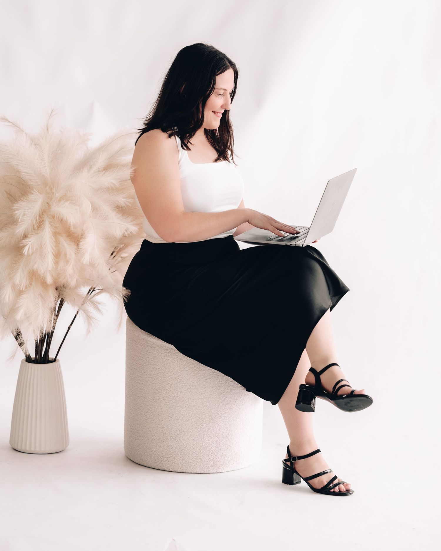 A smiling woman in a white top and black skirt sits on a stool typing on a laptop beside a vase of pampas grass.