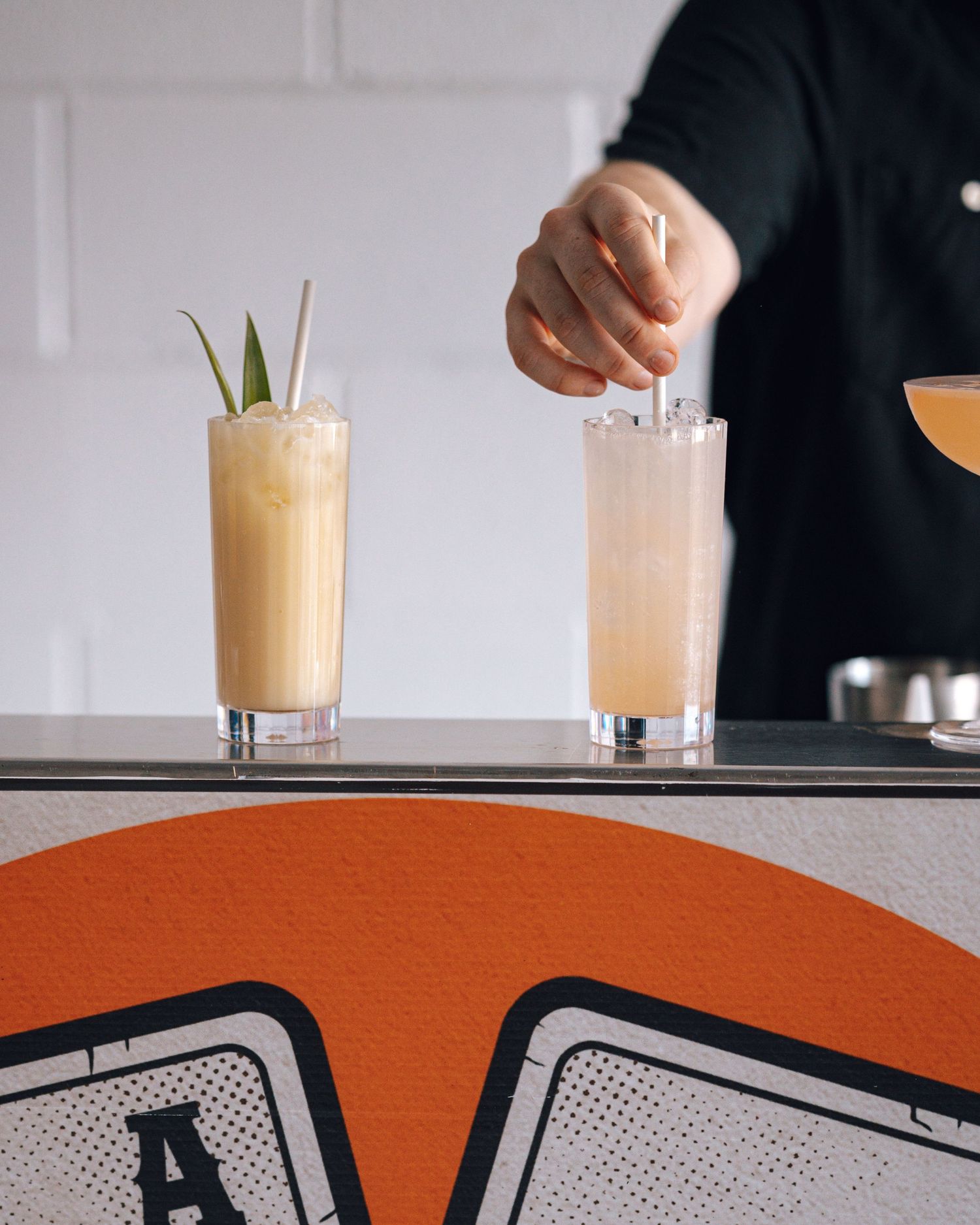 Bartender stirs a cocktail beside a creamy tropical drink garnished with pineapple leaves on a Pocket Ace Bartending mobile bar.