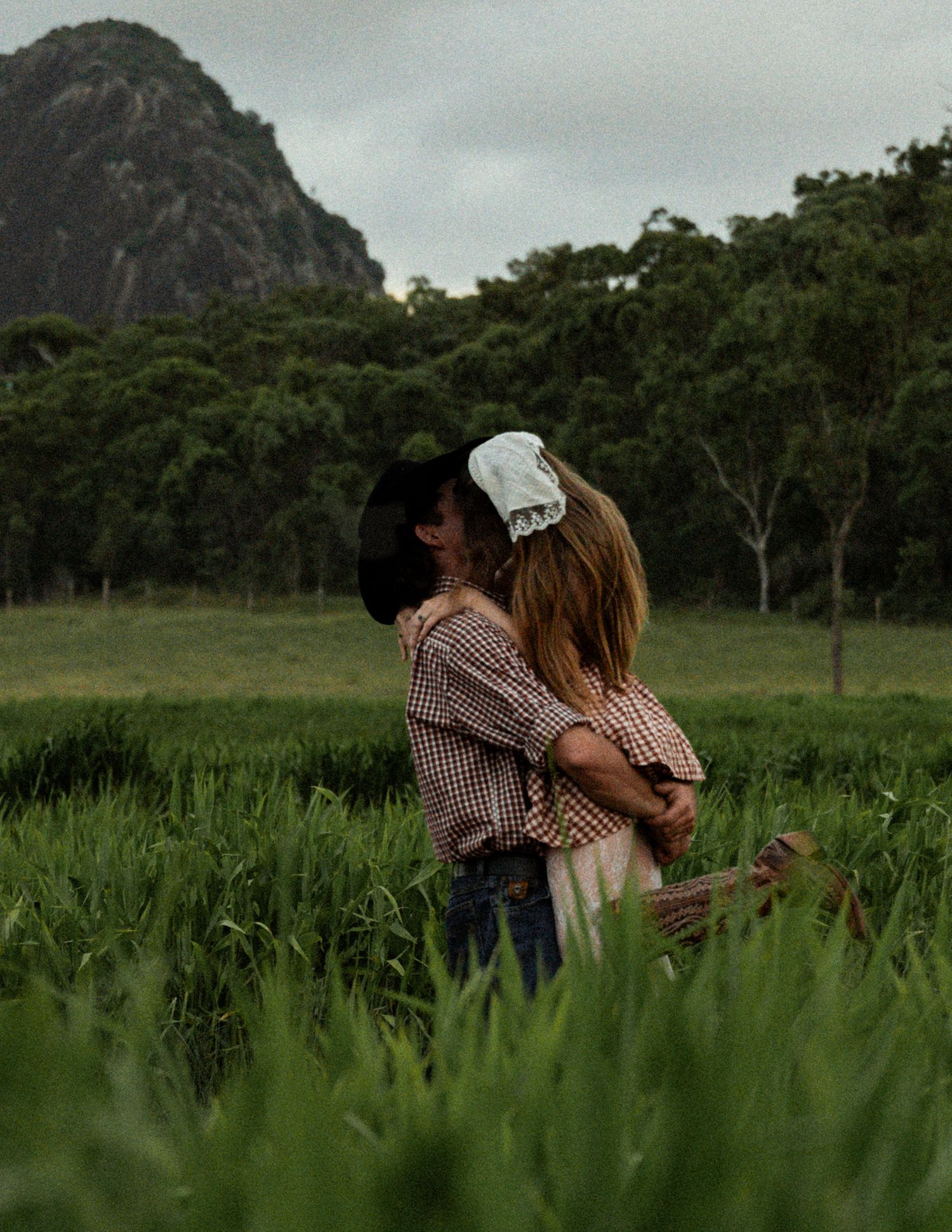 Couple in country-style clothing embracing in a lush green grass field with a rocky mountain and trees in the background.