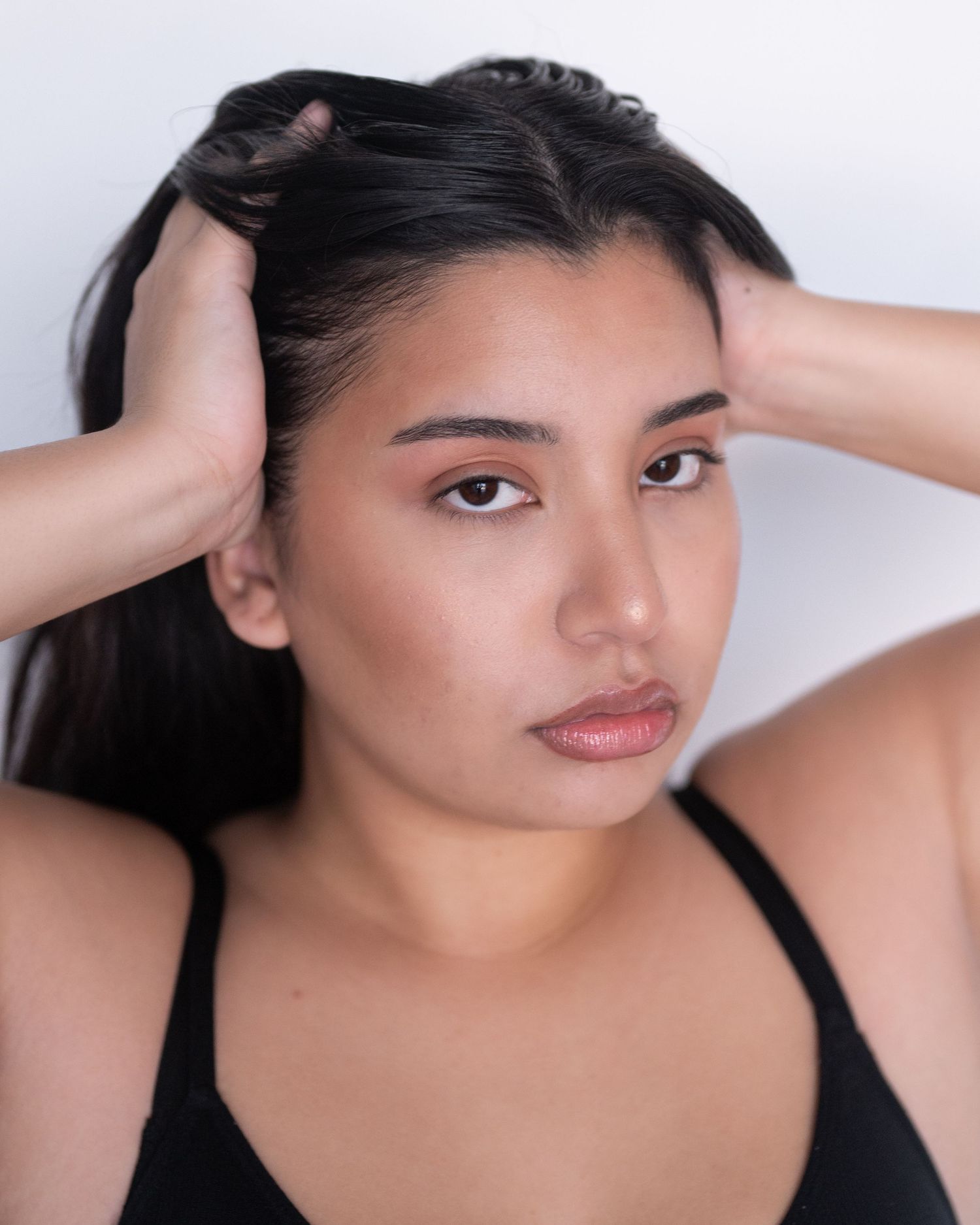 Close-up of a woman with dark hair pulled back by both hands, wearing a black top against a soft white background.