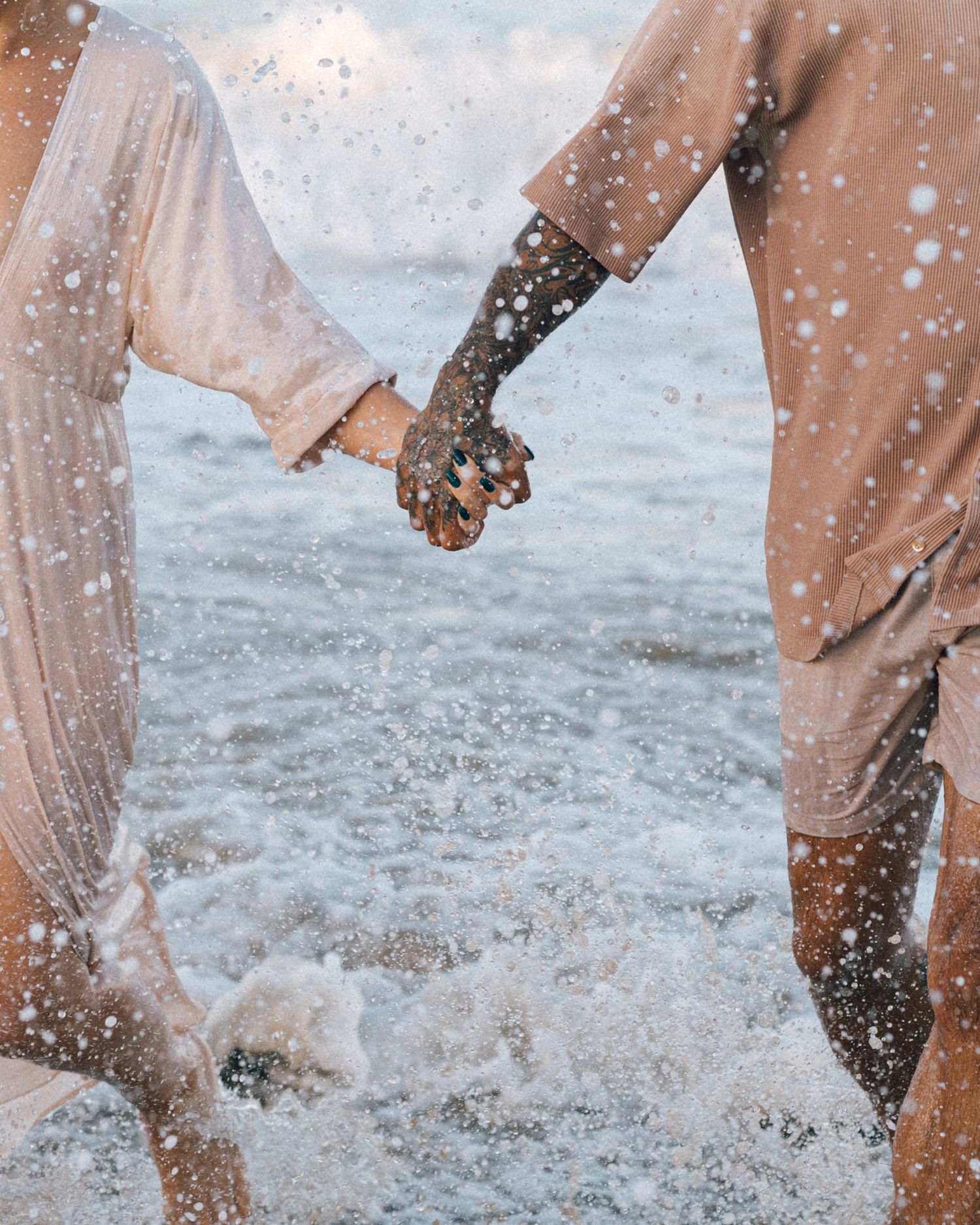 A couple in neutral linen outfits holds hands while walking through splashing ocean waves at the beach.