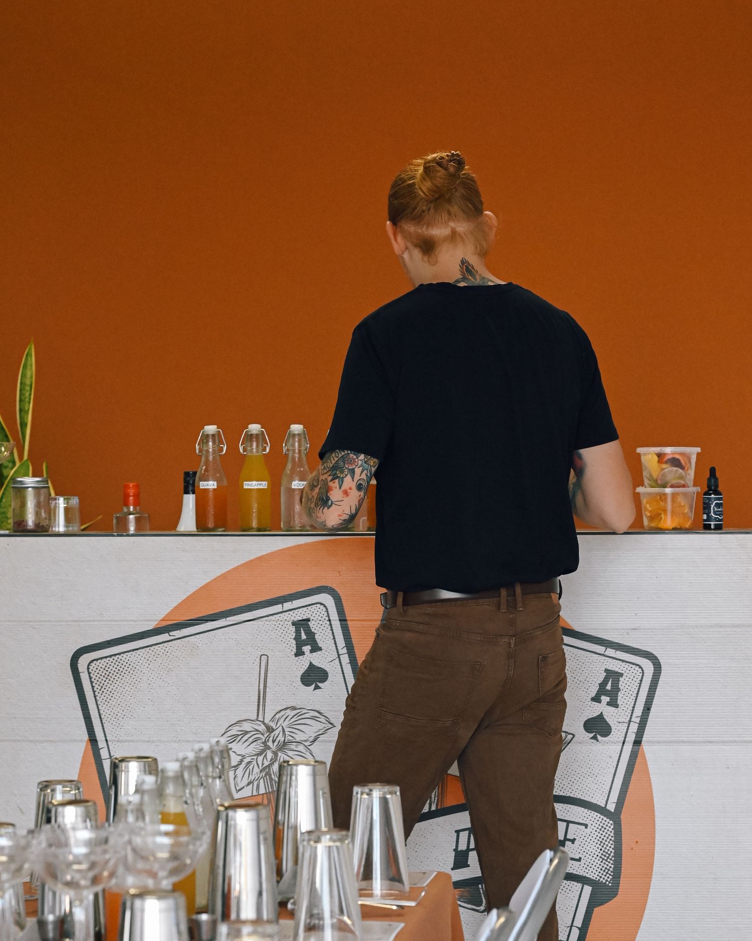 Bartender in a black t-shirt with arm tattoos works behind a white bar with a playing card design against a burnt orange wall.
