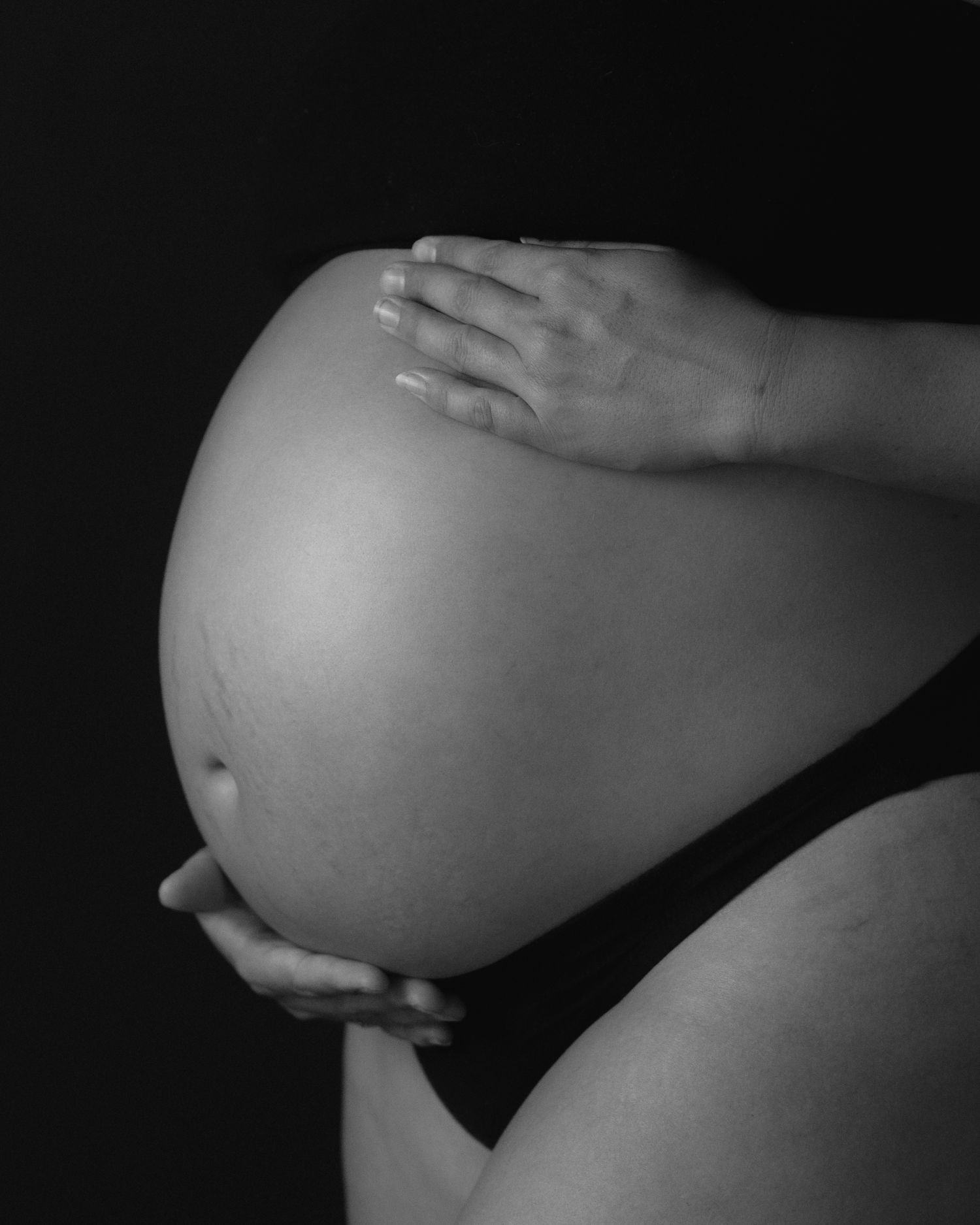 Black and white side profile of a large pregnant belly with hands resting gently on top against a dark background.