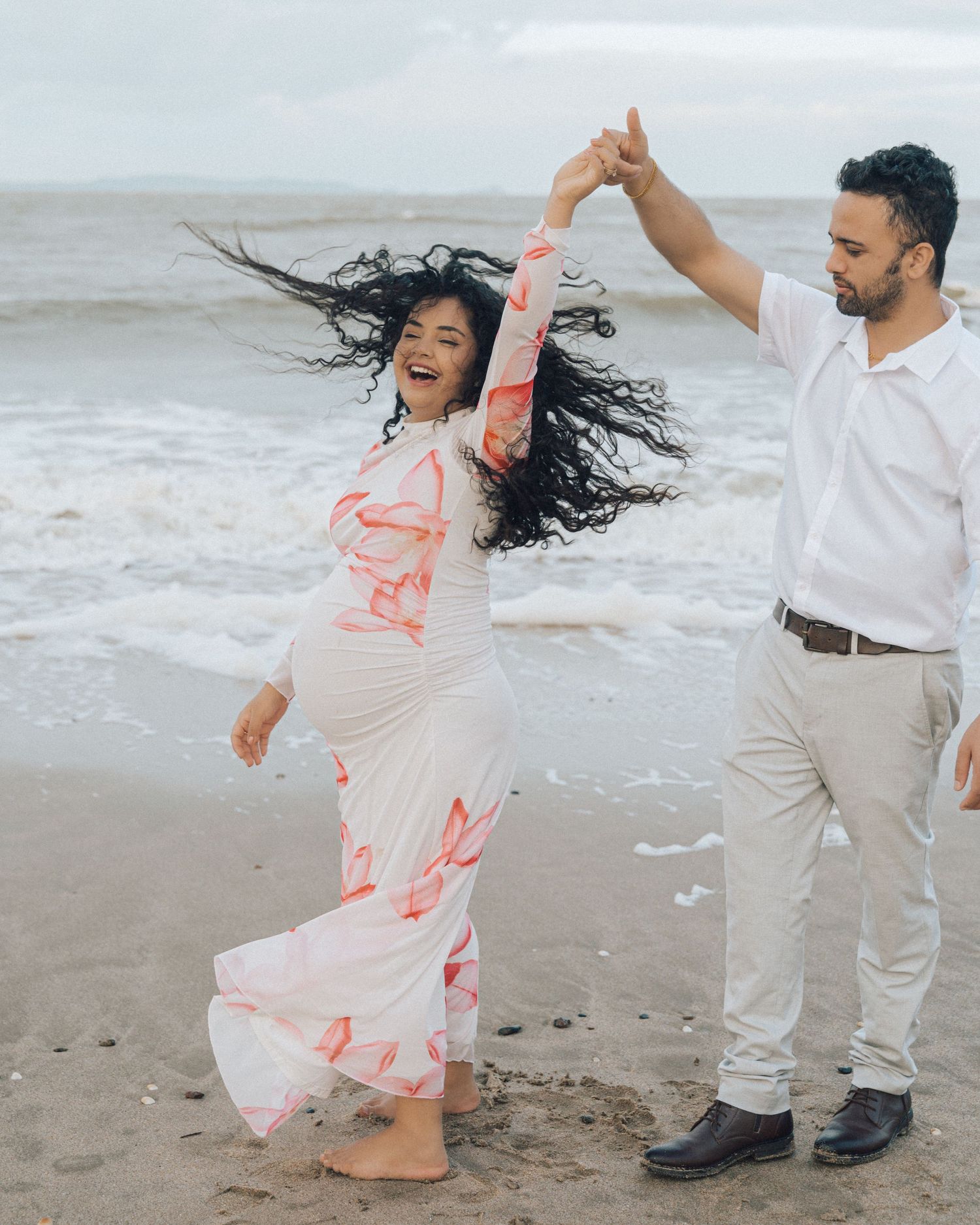 Joyful pregnant woman with flowing hair spins barefoot on the sand while her partner in a white shirt holds her hand.