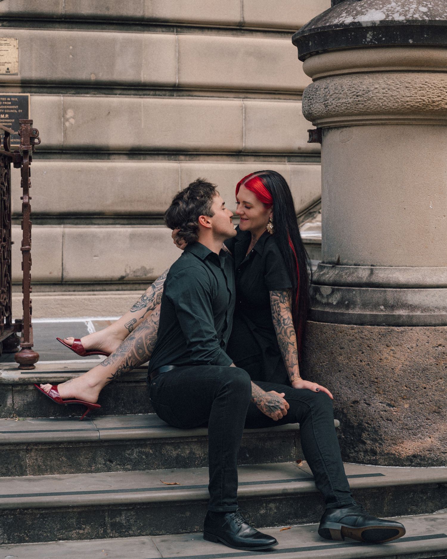 A tattooed couple in black sits close together on sandstone steps, smiling at each other, the woman with red-streaked hair and red heels.
