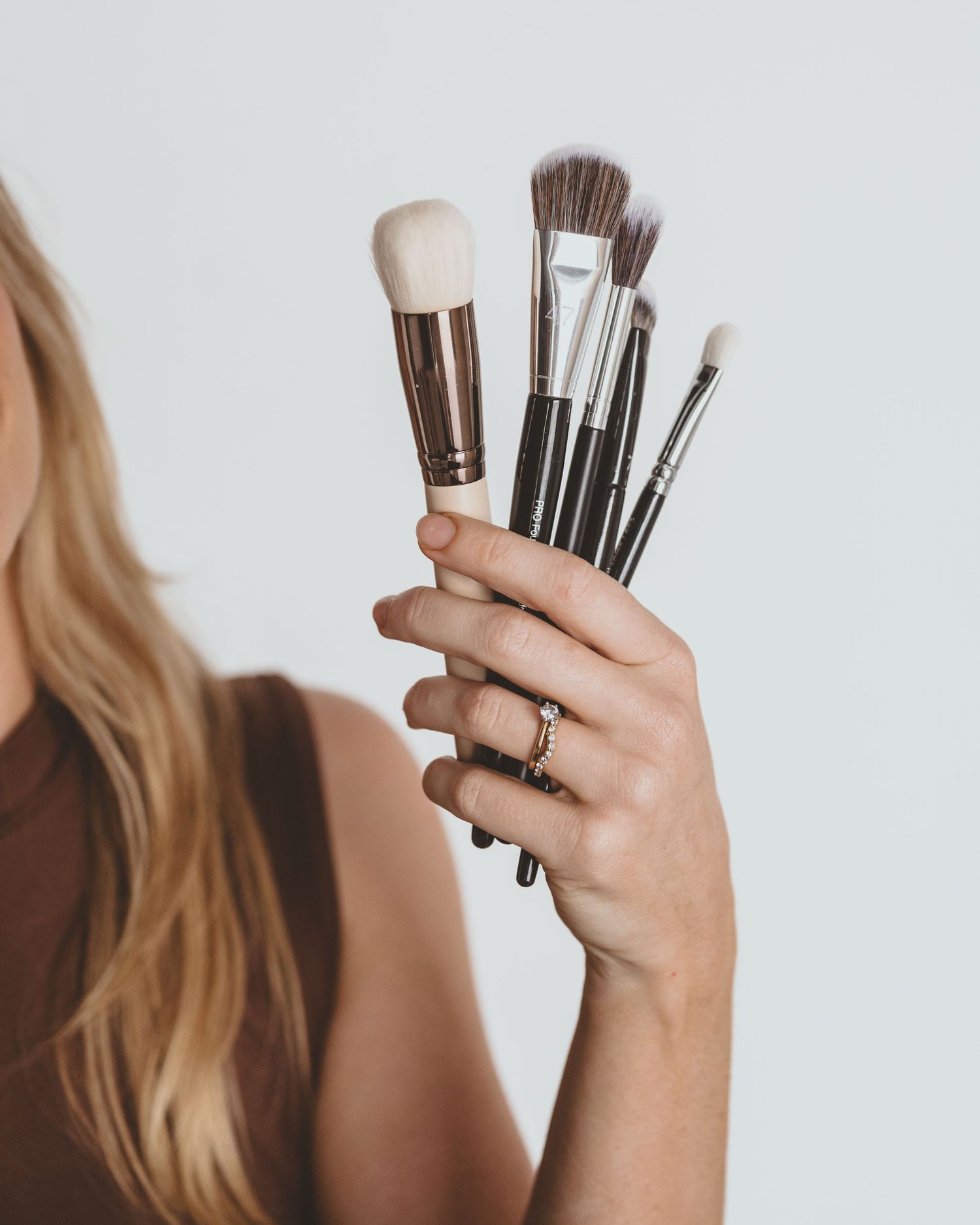 Woman with long blonde hair holds a bunch of makeup brushes of various sizes in her raised hand.
