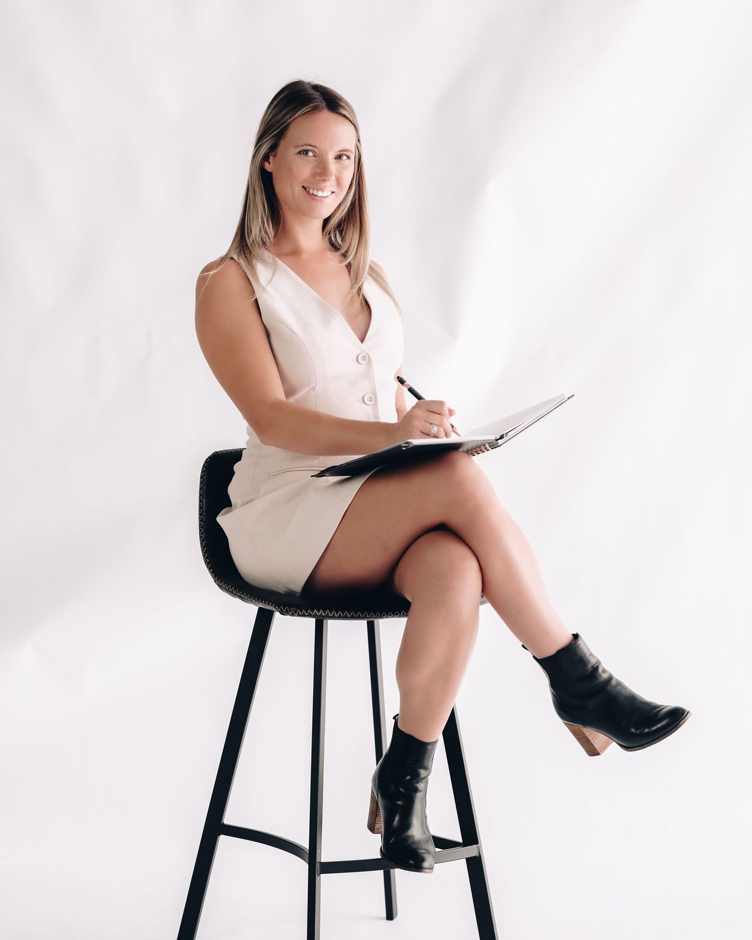 A smiling woman in a cream vest dress sits on a black stool, writing in a notebook in a bright studio.