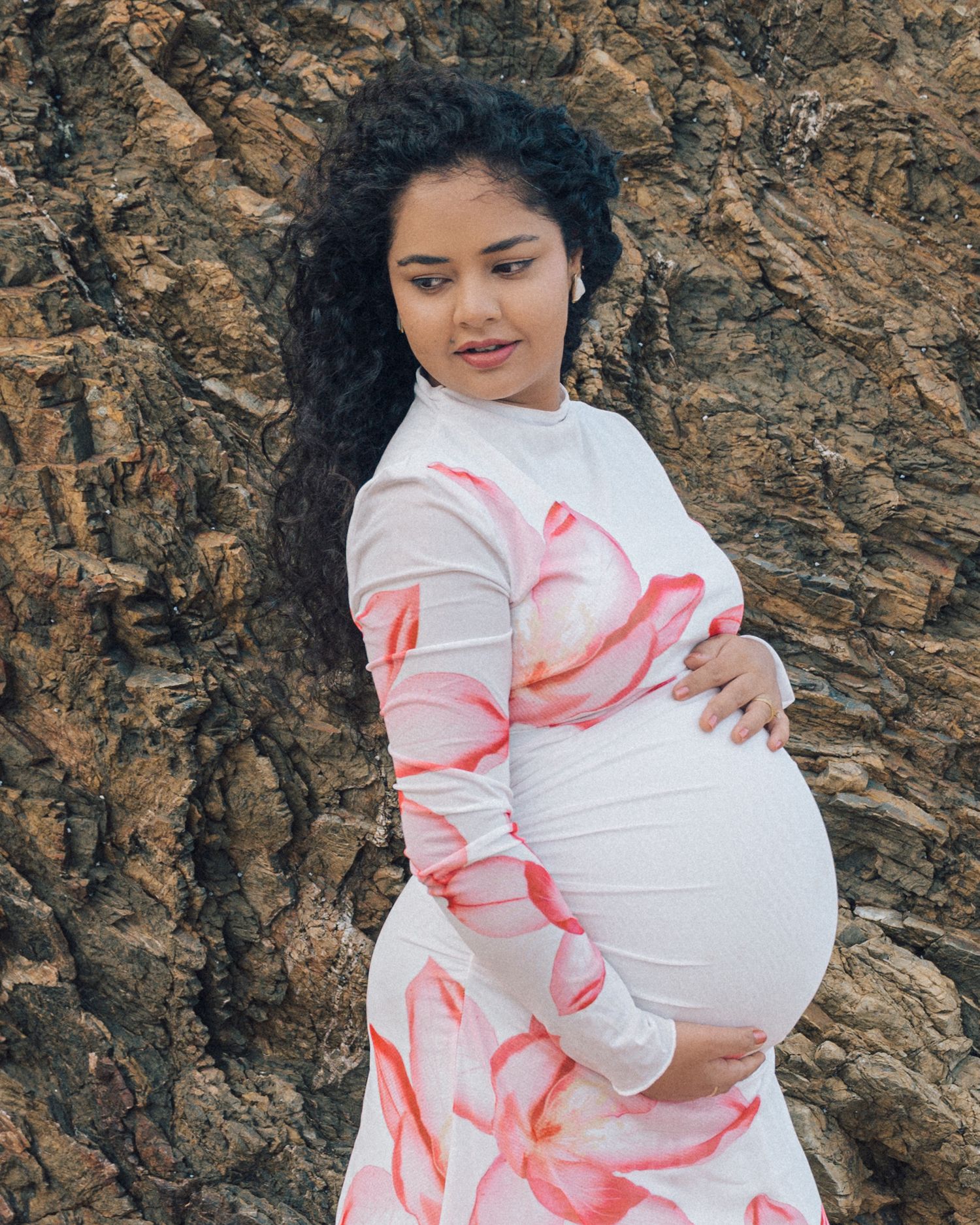 Pregnant woman with long curly dark hair in a pink floral fitted dress poses beside a rugged rock wall.