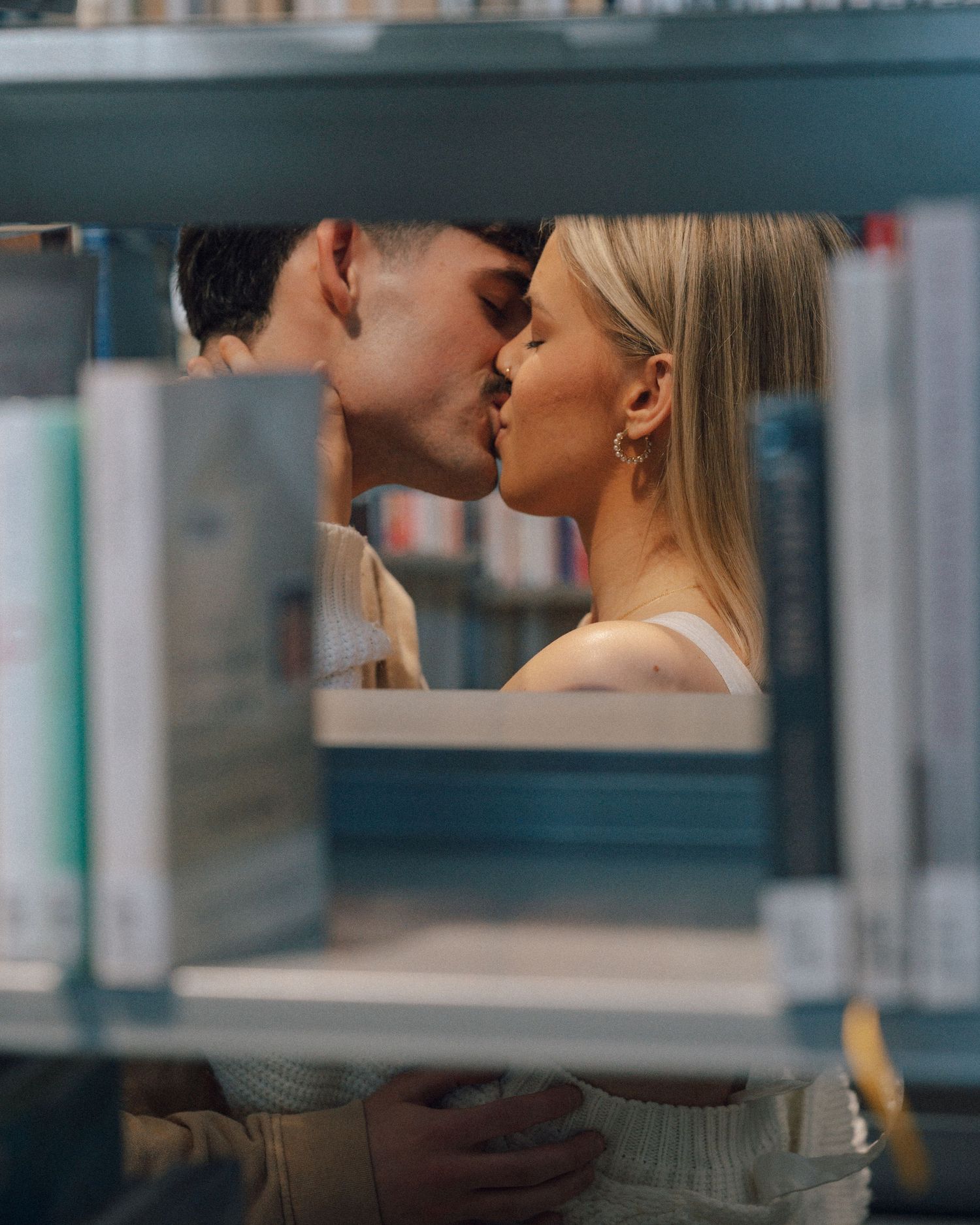 Couple sharing a romantic kiss seen through a gap in library bookshelves, framed by rows of books.