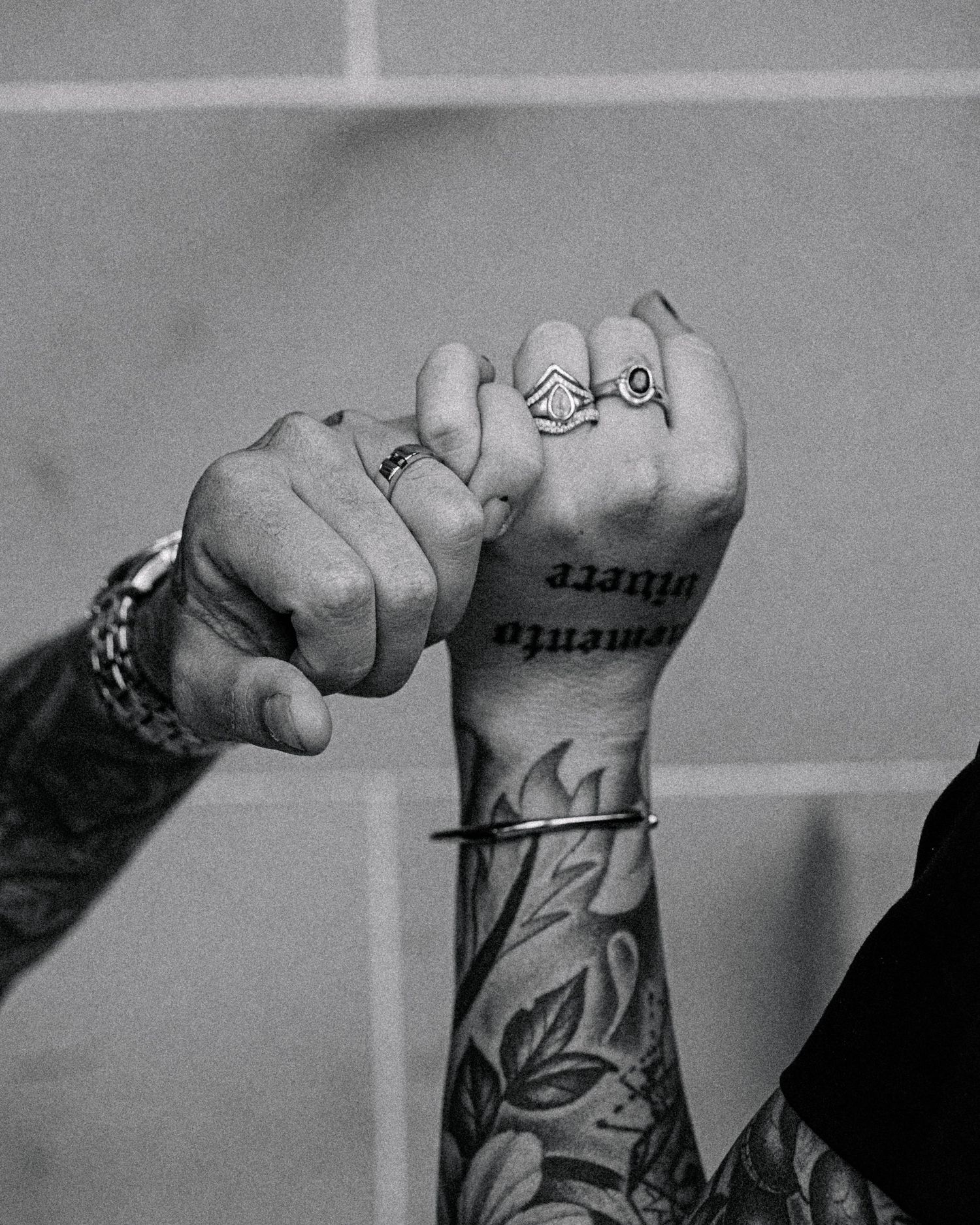Black and white close-up of two hands with linked pinkies, stacked rings, a cuff bracelet and a floral forearm tattoo with Latin script.