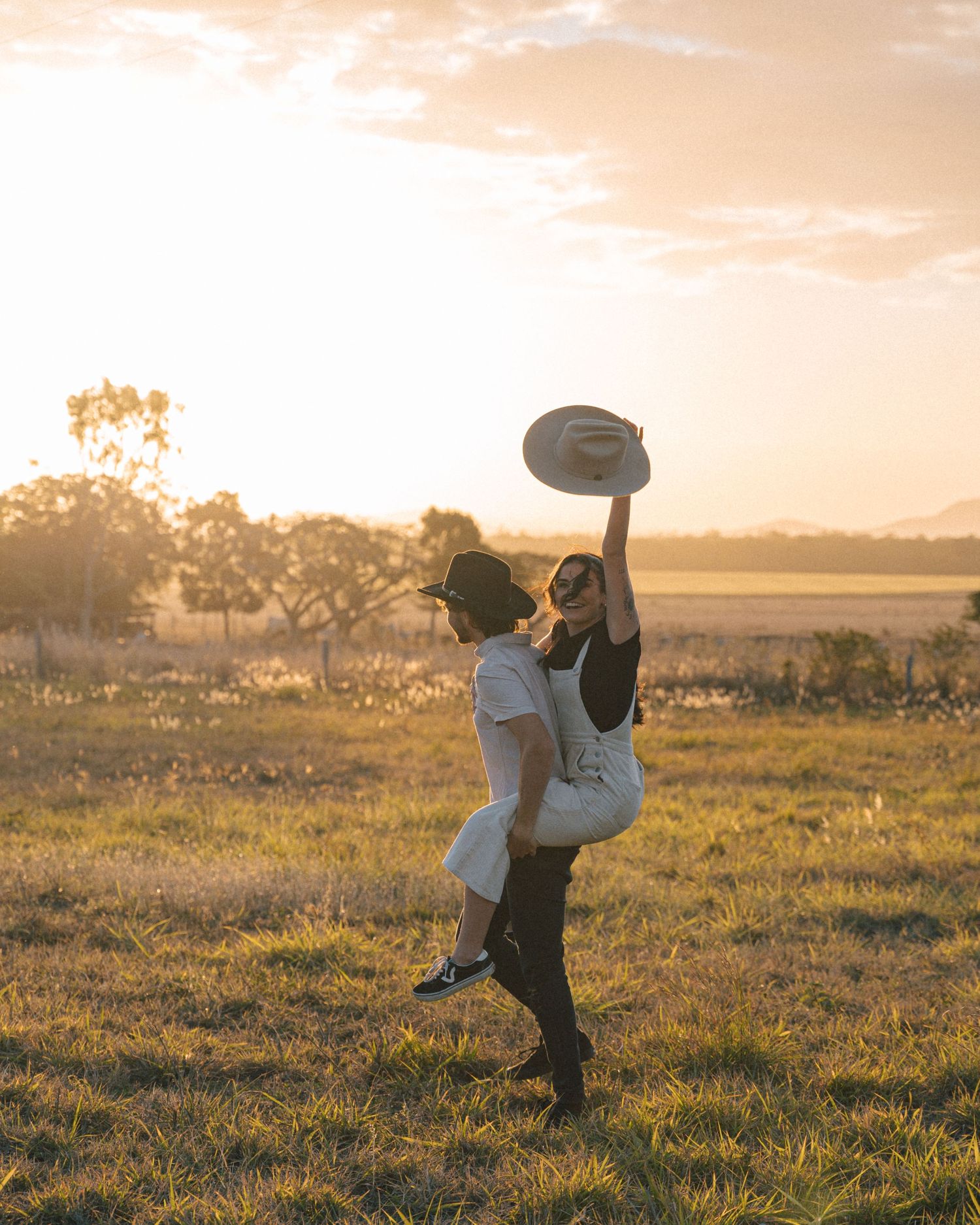 Woman in a white dress lifted joyfully by a man in a black hat, holding her hat high in a sunset field.