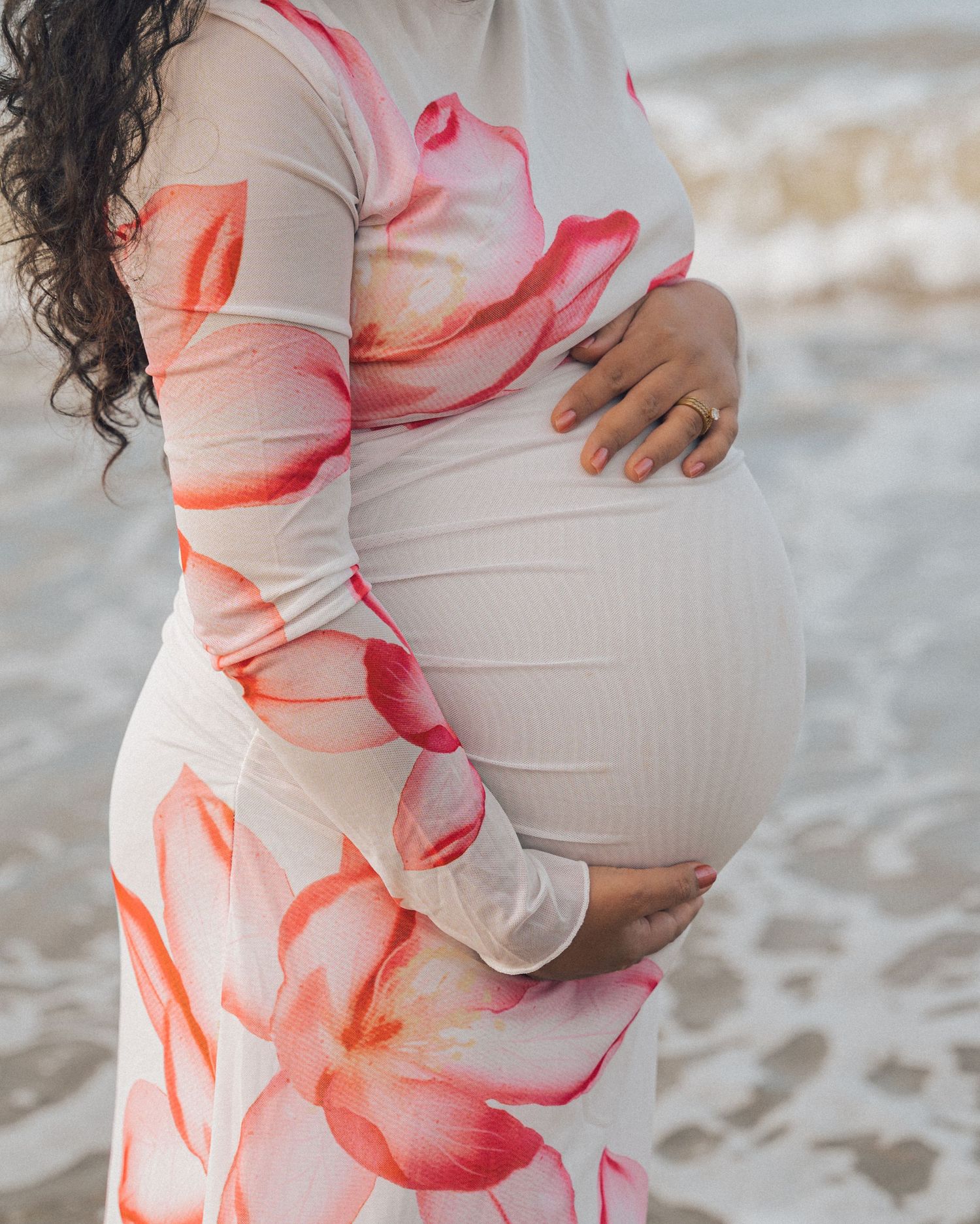 Side view of a pregnant woman in a pink floral dress holding her round belly with soft ocean waves in the background.