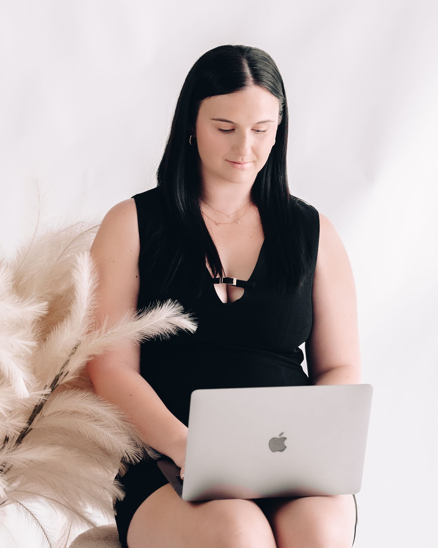 Woman with dark hair in a black sleeveless top works on a silver laptop seated beside dried pampas grass.