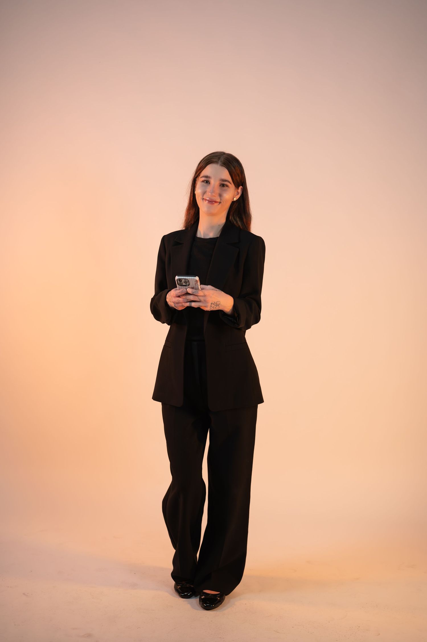 Woman in a black blazer and trousers stands smiling against a warm peach backdrop while holding a smartphone.