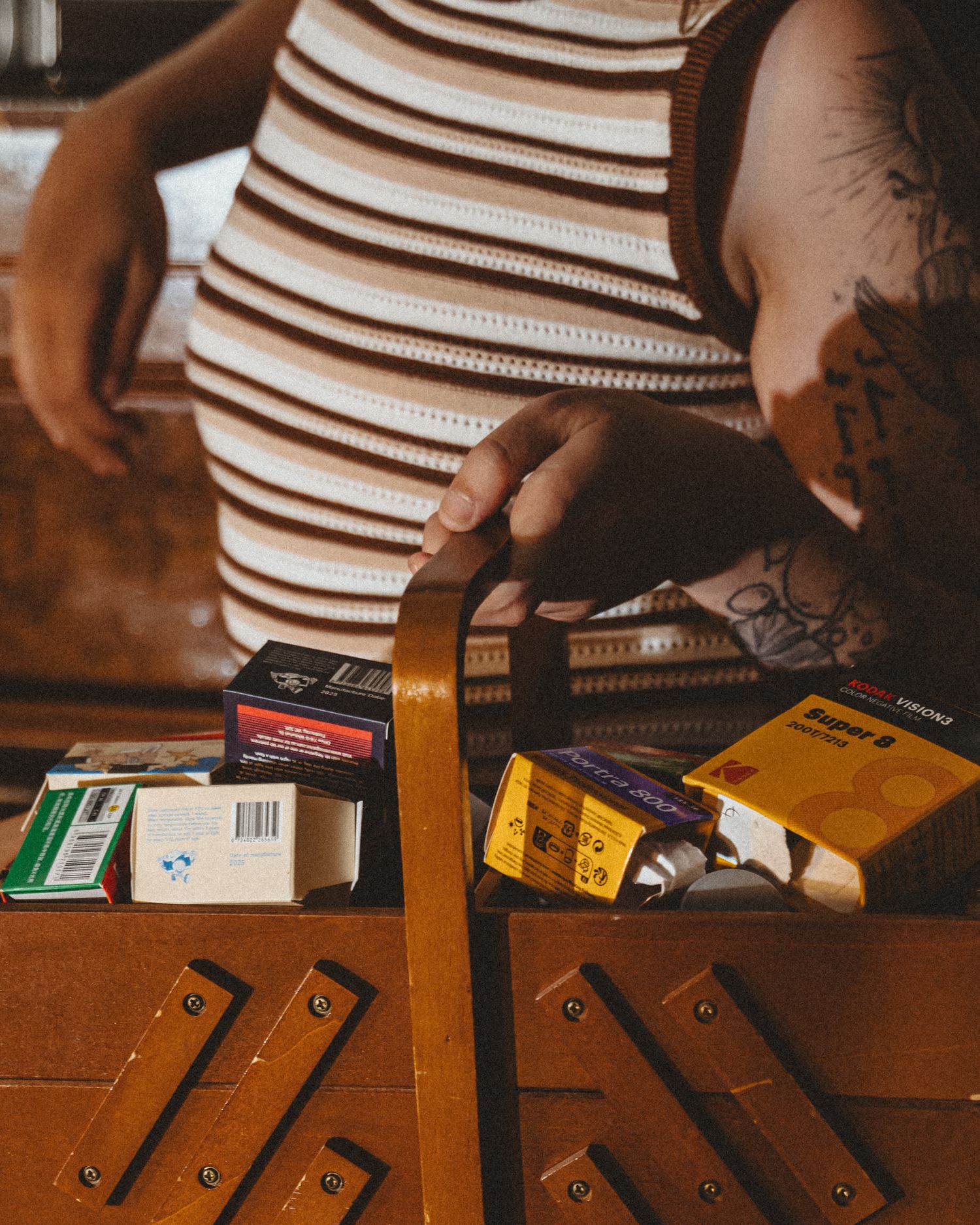 Close-up of a wooden vintage sewing box filled with colorful film boxes, held by a tattooed person in a brown striped top.