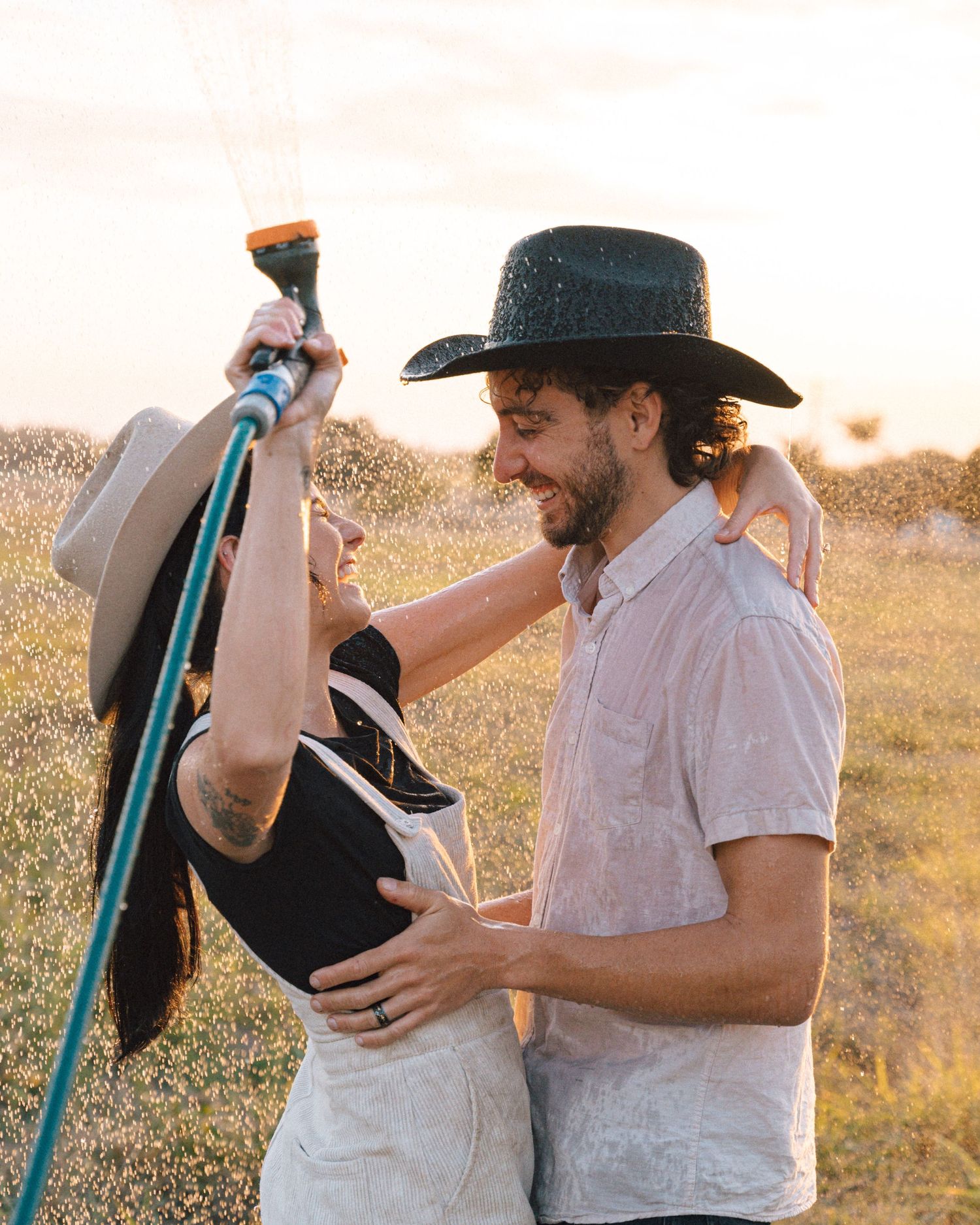 Smiling couple in cowboy hats embracing in a sunlit field while the woman sprays a garden hose overhead.
