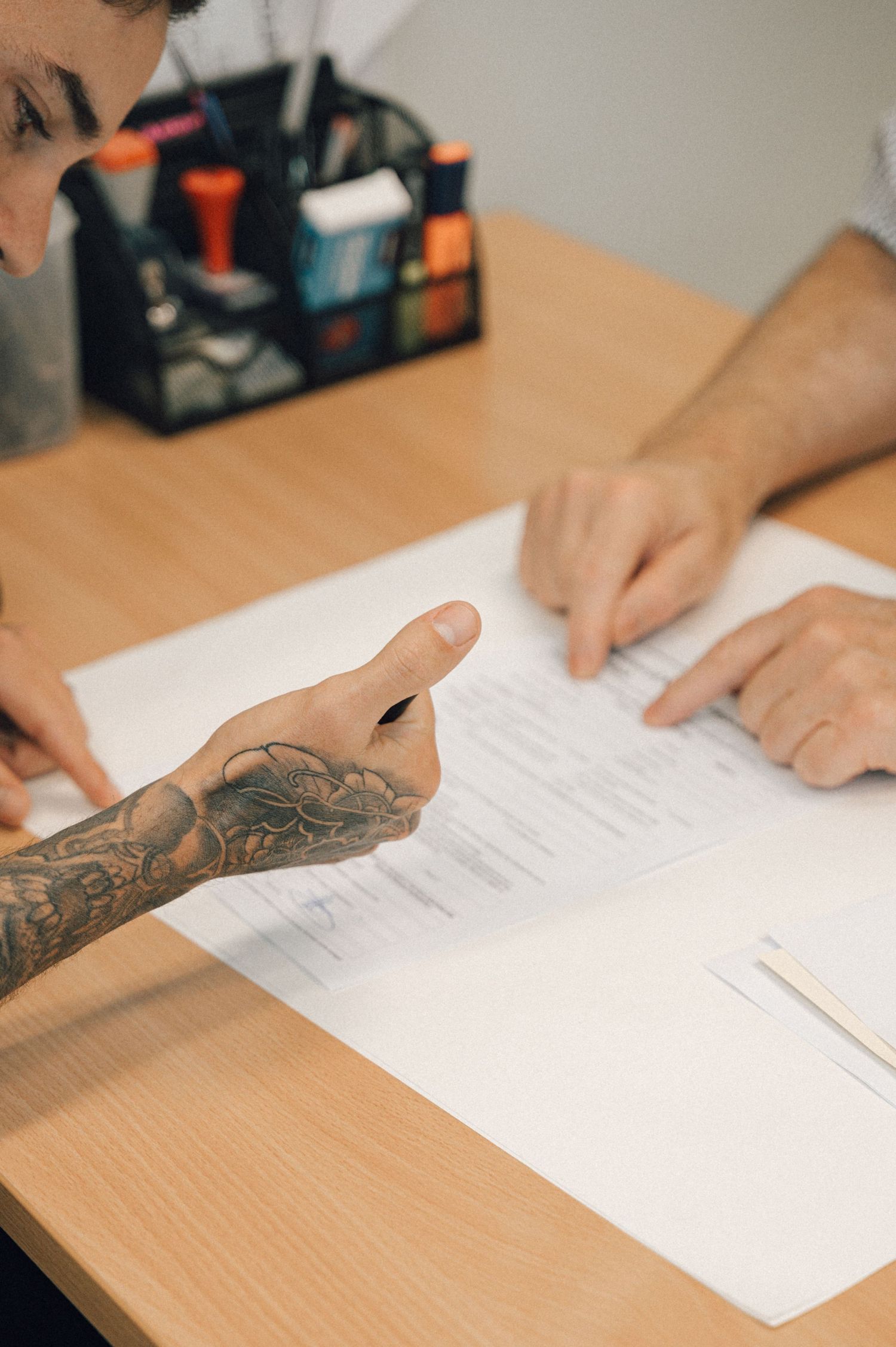 Tattooed hand holding a pen over marriage paperwork on a wooden desk with a stationery organizer.
