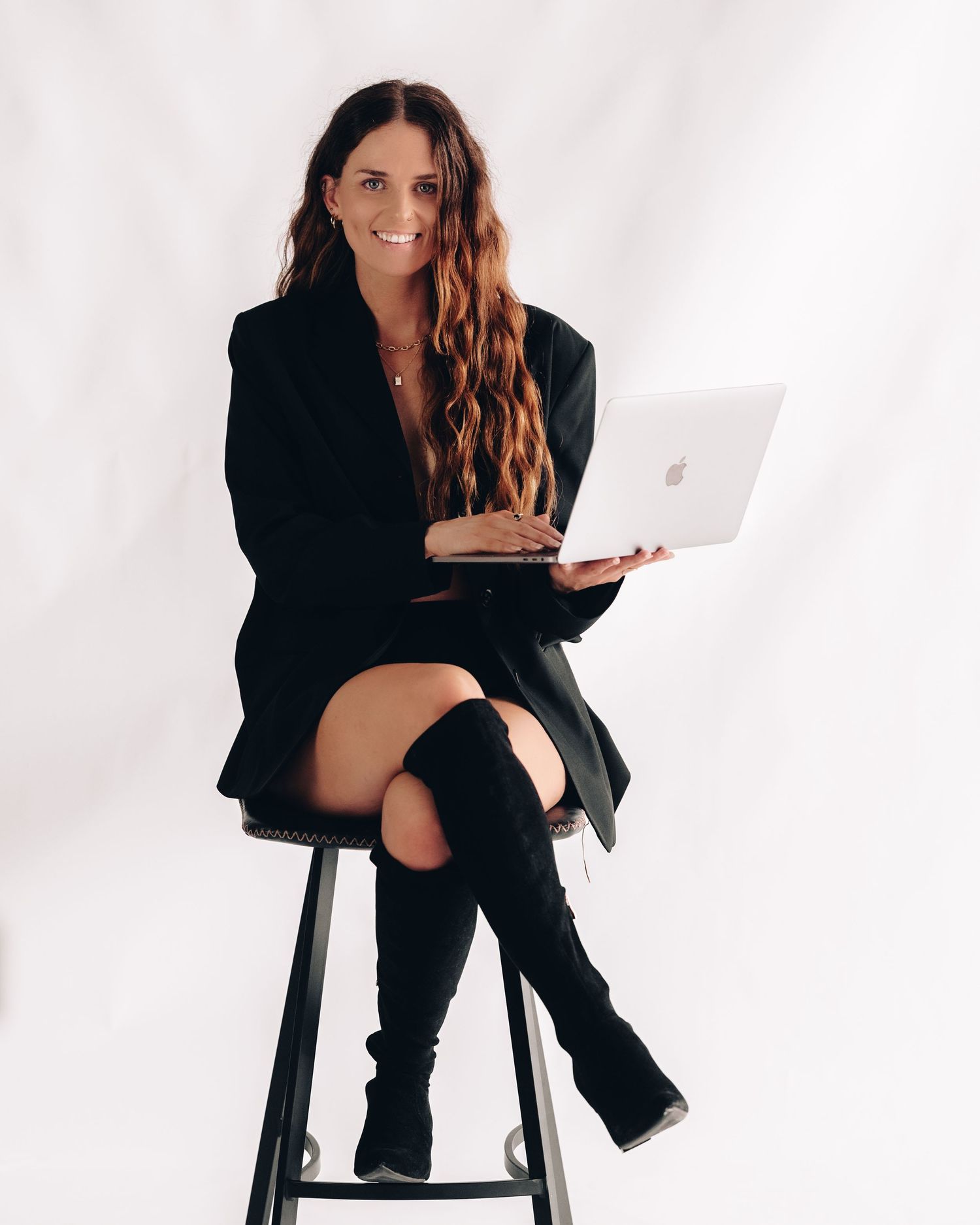 Smiling woman in a black blazer and knee-high black boots sits on a stool holding an open laptop against a white backdrop.