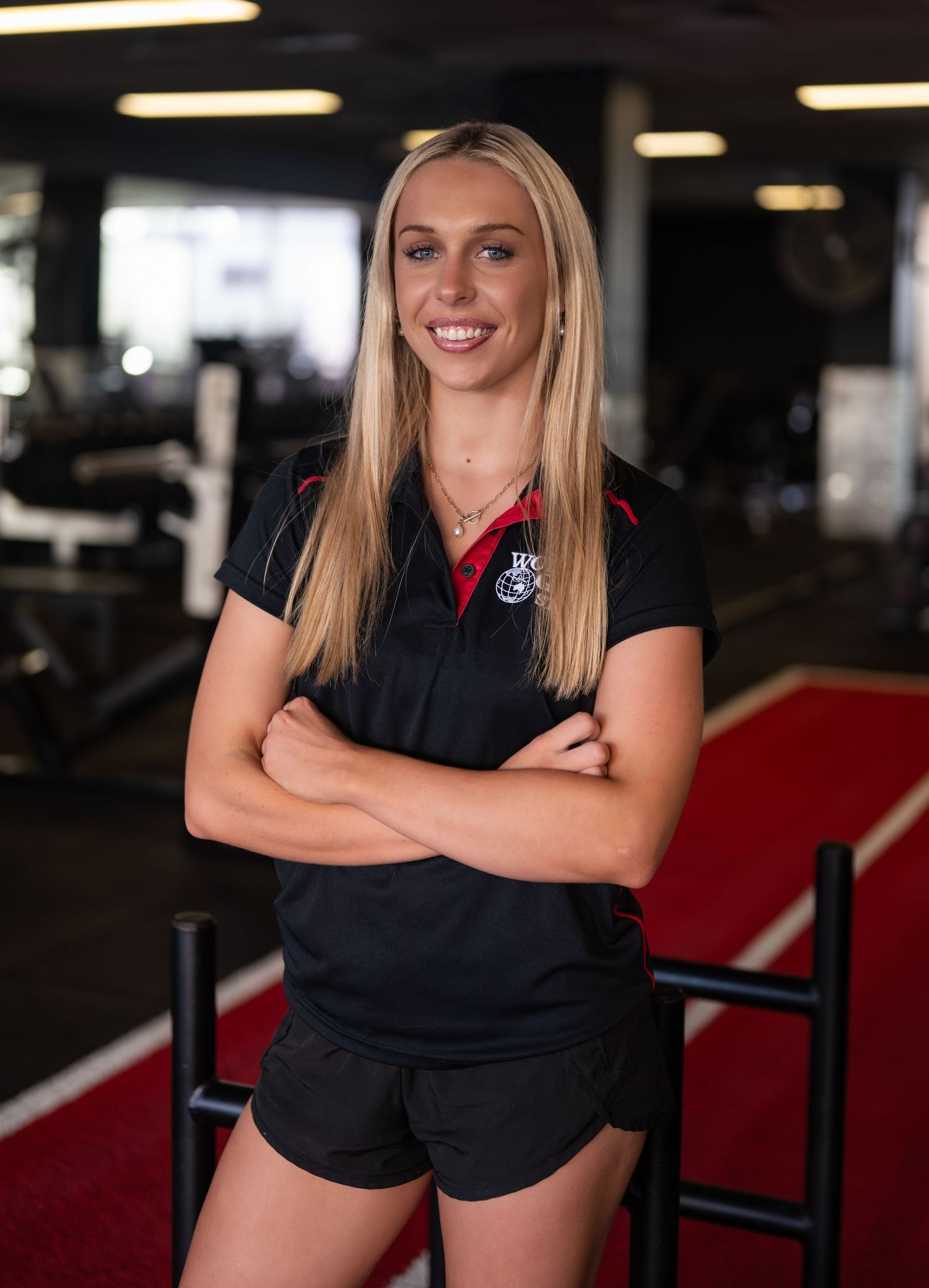 Smiling woman with long blonde hair and arms crossed stands in a gym wearing a black staff polo and shorts.