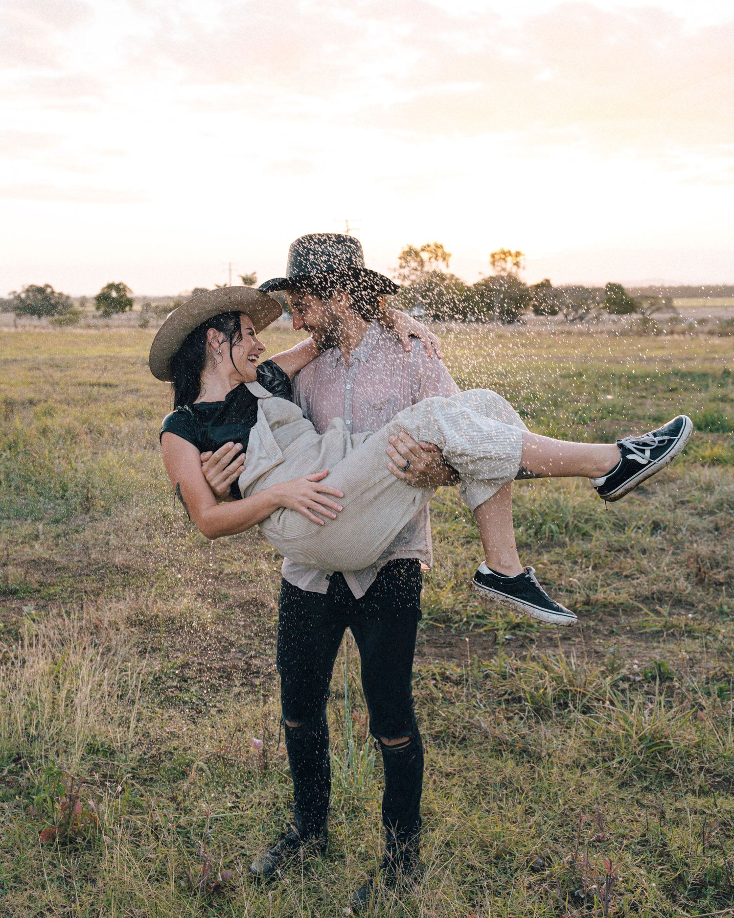 A man in a black hat lifts a laughing woman in overalls and sneakers in his arms in a golden grassy field.