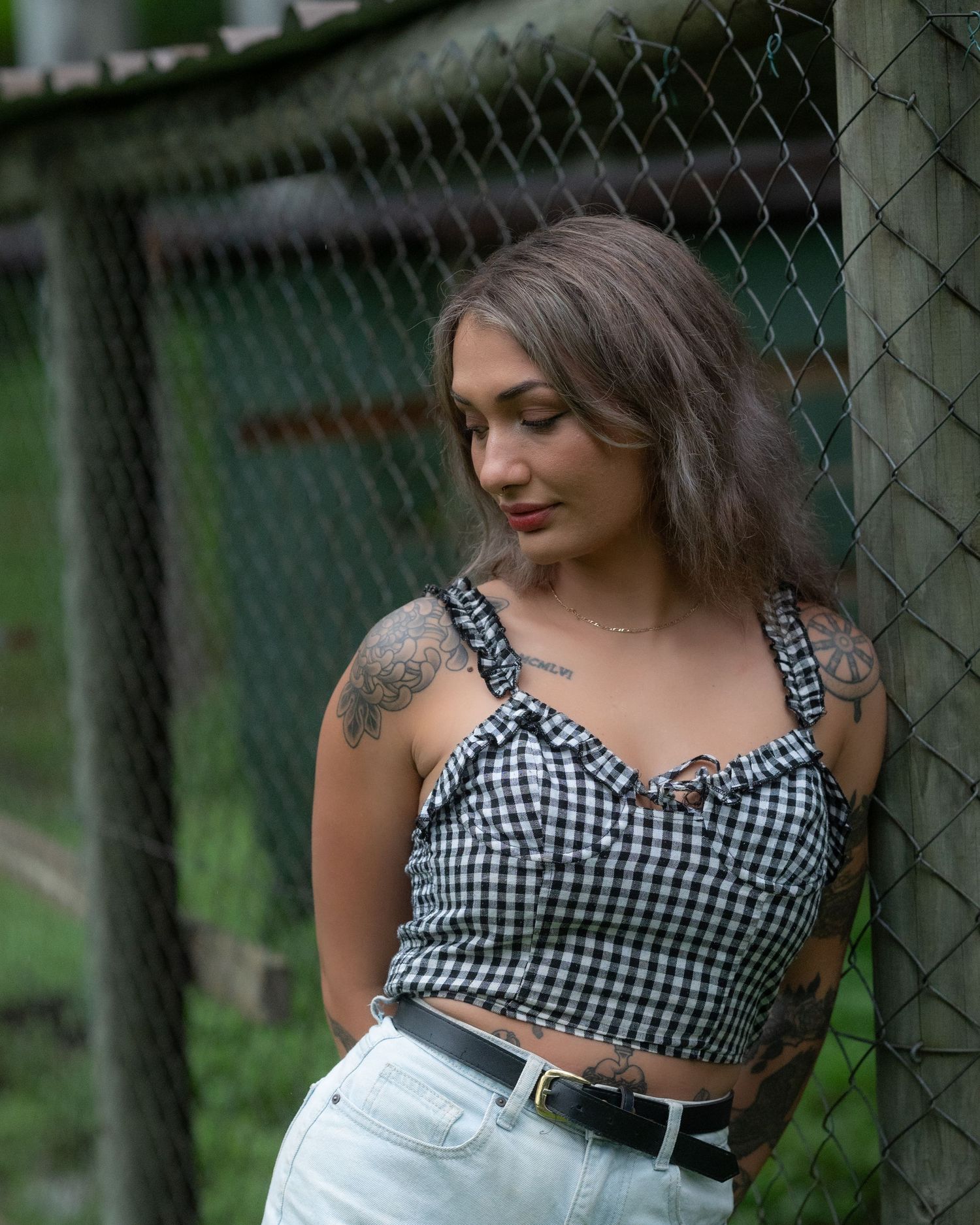 Tattooed woman in a gingham top and high-waisted shorts gazes downward while leaning on a chain-link fence post.