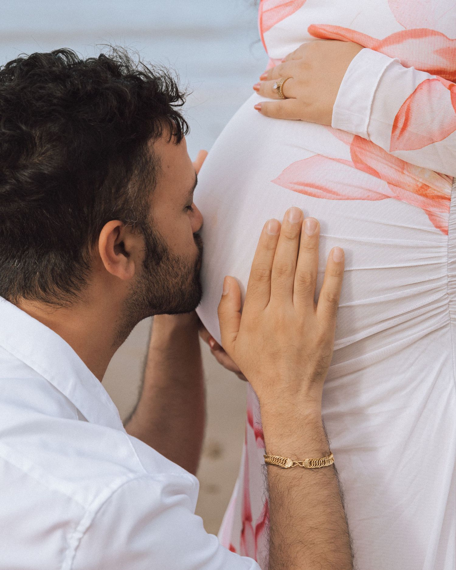 Man in white shirt kisses his pregnant partner's belly on the beach, wearing a gold bracelet during a maternity shoot.