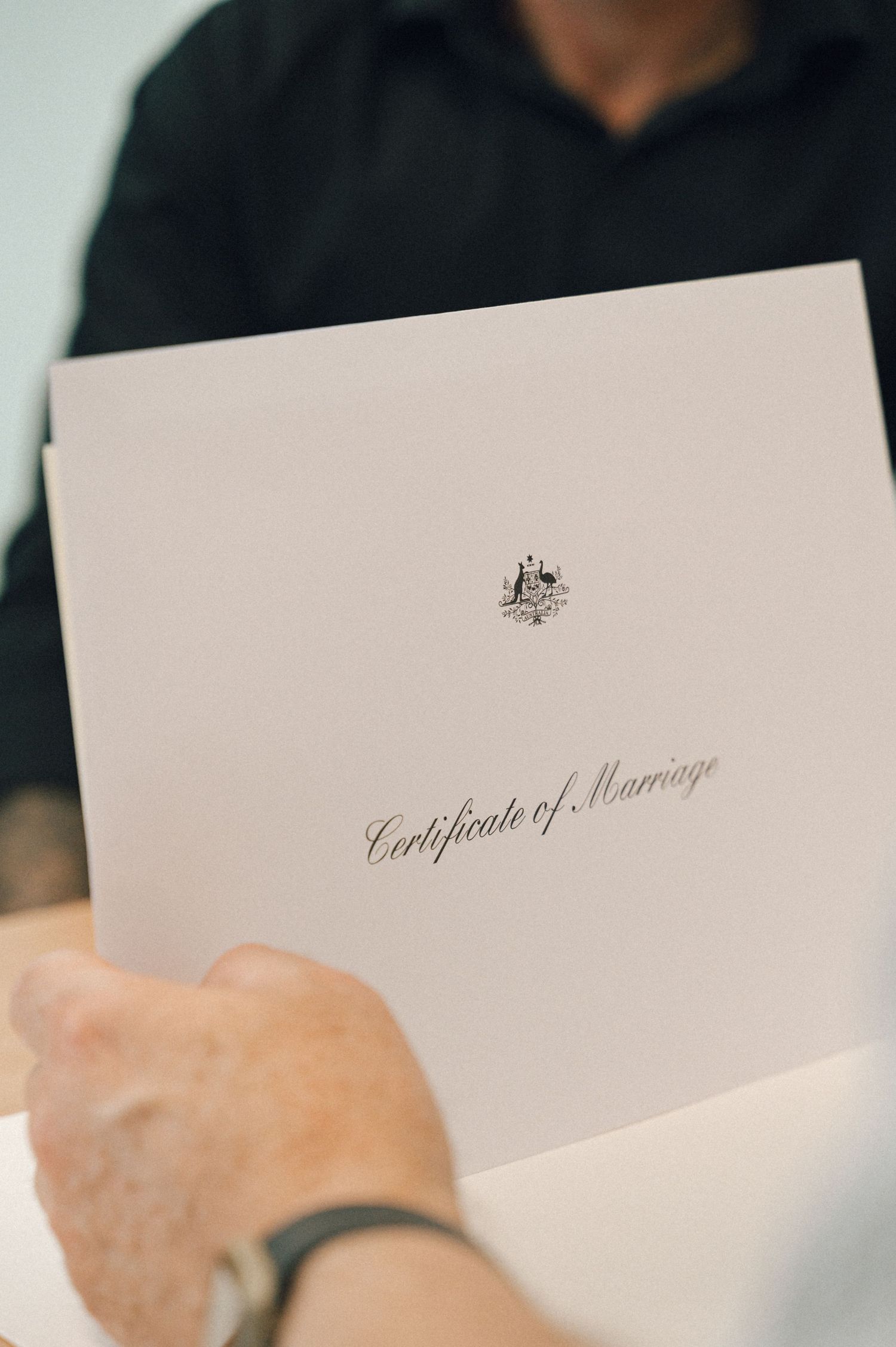 Hand holding a white Australian Certificate of Marriage folder with an embossed crest.