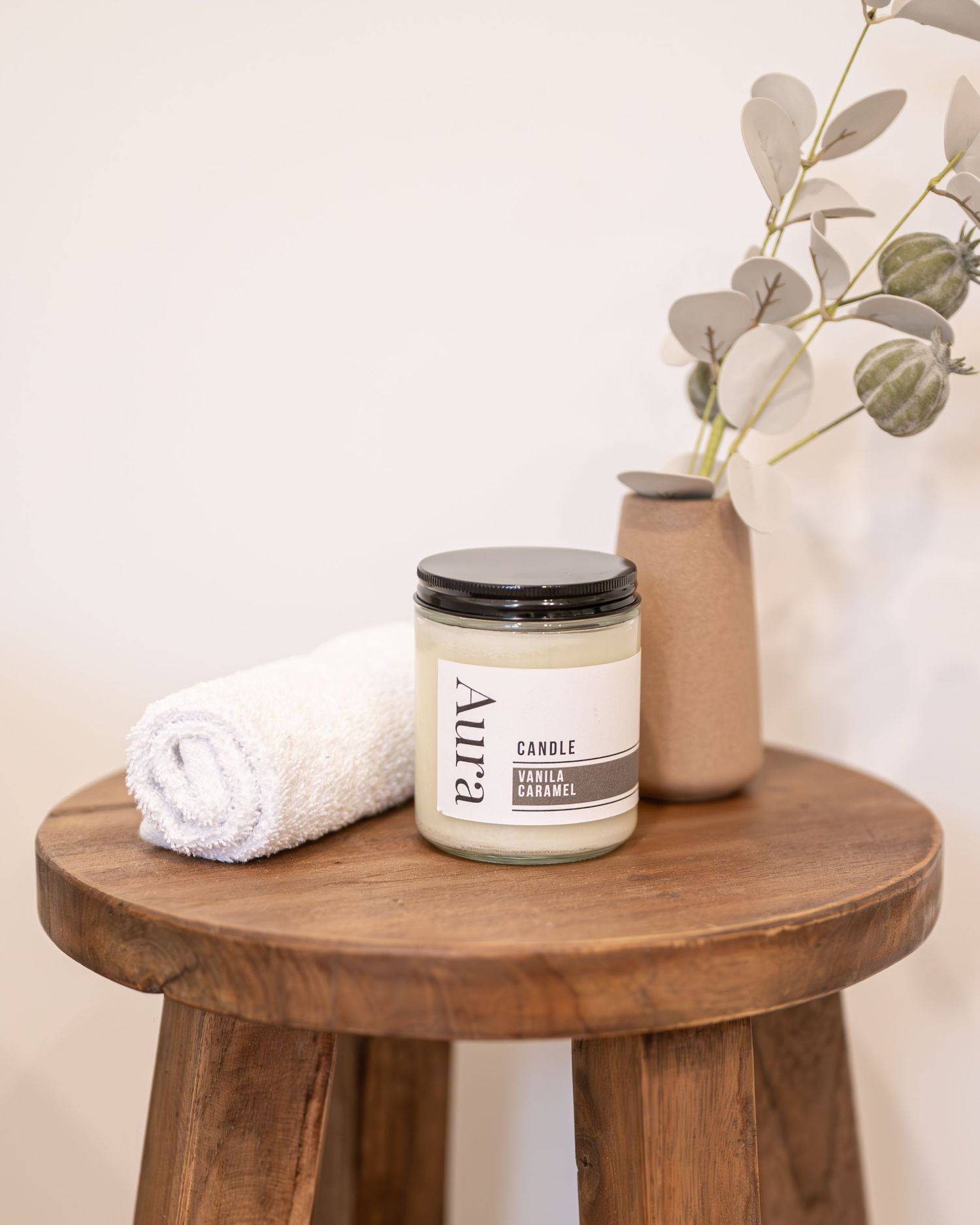 Aura vanilla caramel candle in a glass jar on a wooden stool beside a rolled white towel and a vase with eucalyptus stems.