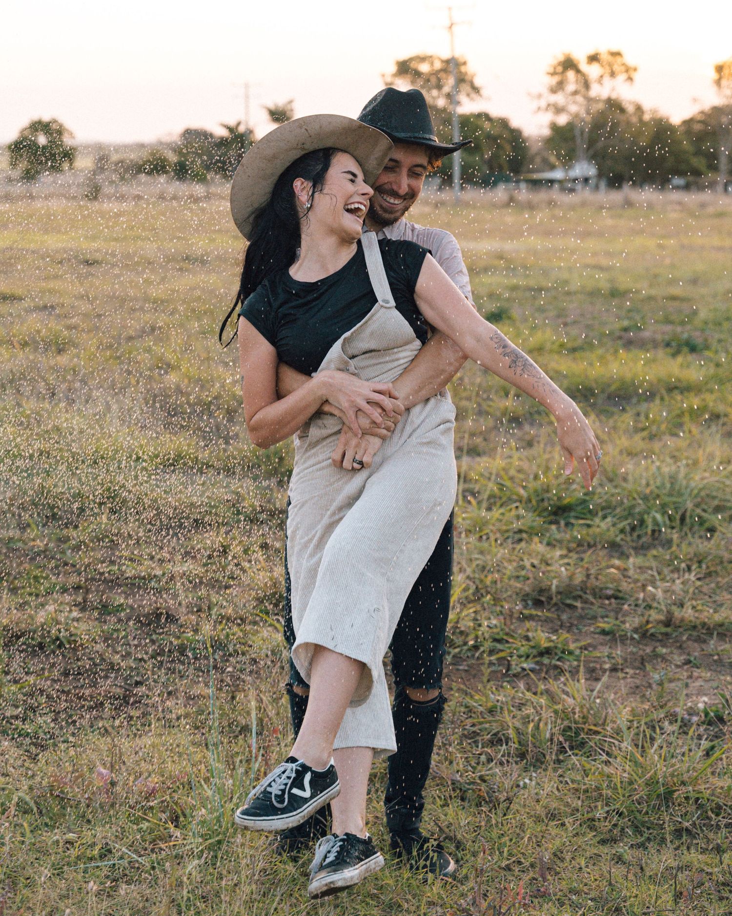 A laughing woman in a cowboy hat and overalls is hugged from behind by a man in a black hat in a field.