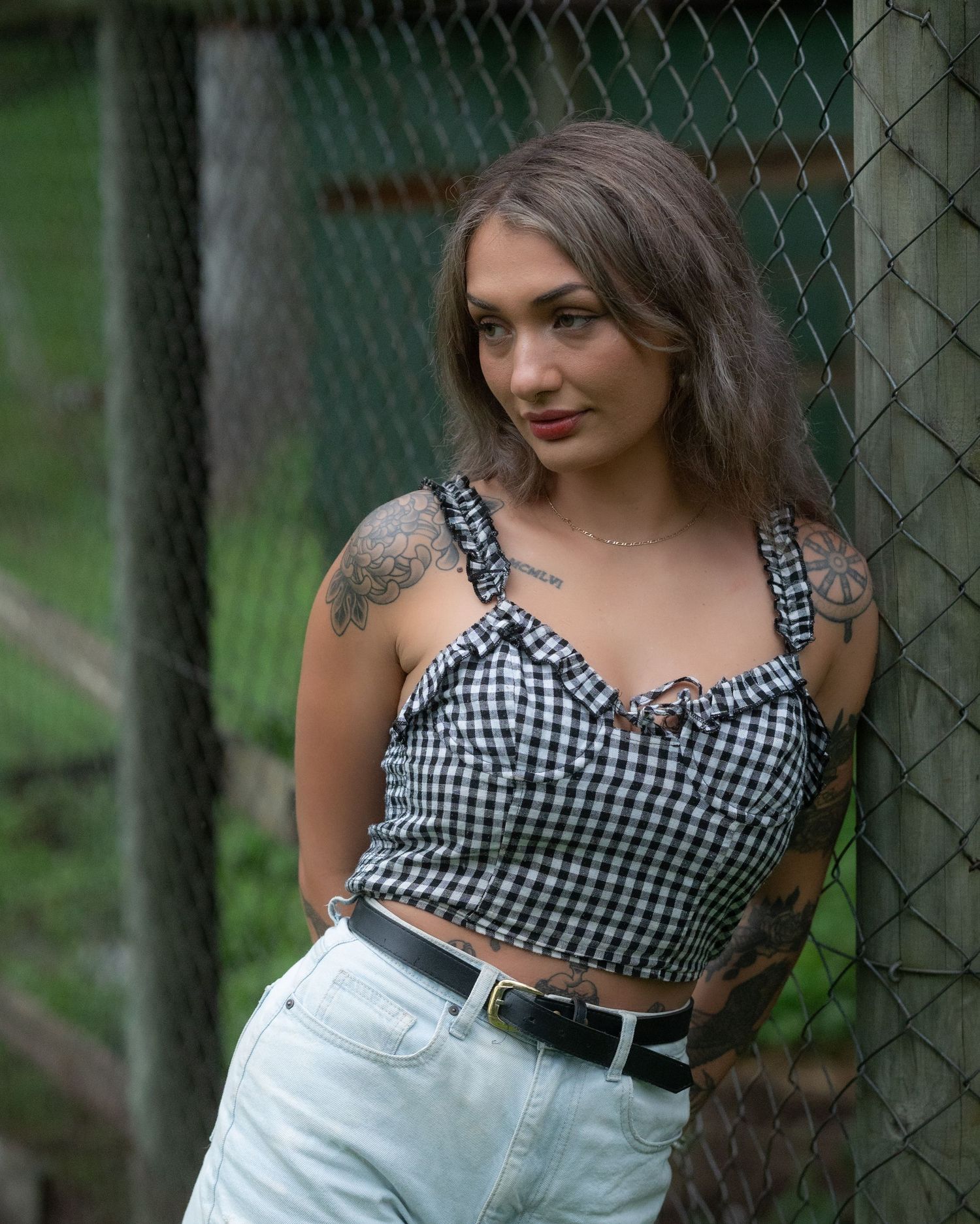 Portrait of a tattooed woman in a black gingham corset top and denim shorts leaning against a chain-link fence outdoors.