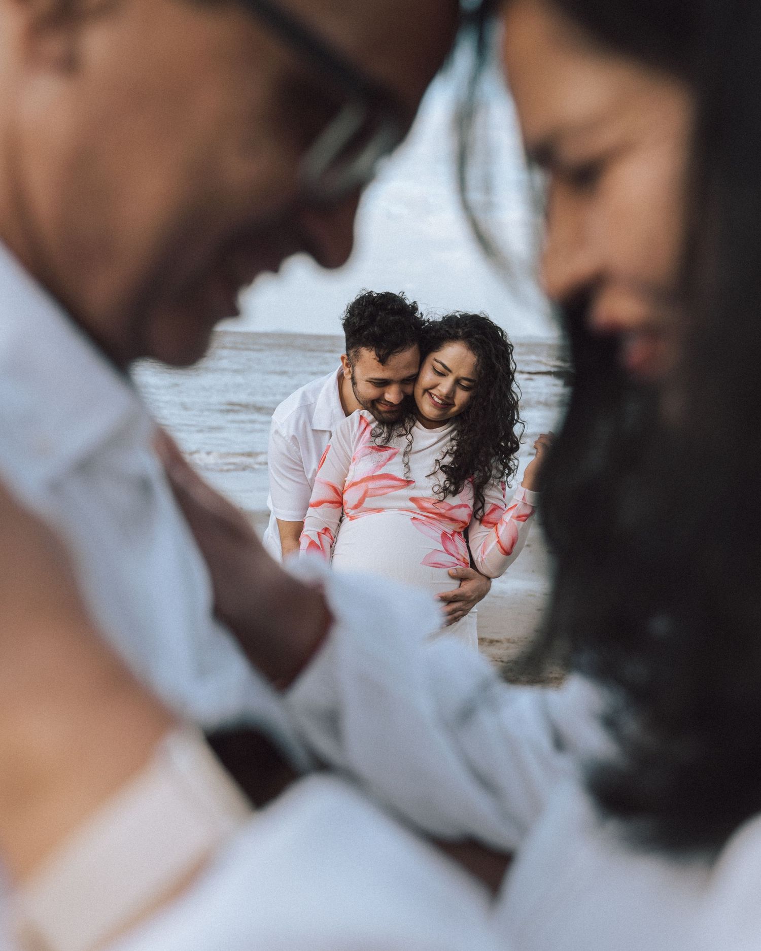 Pregnant couple laughing together on the beach framed through two blurred foreground figures in a creative maternity photo.