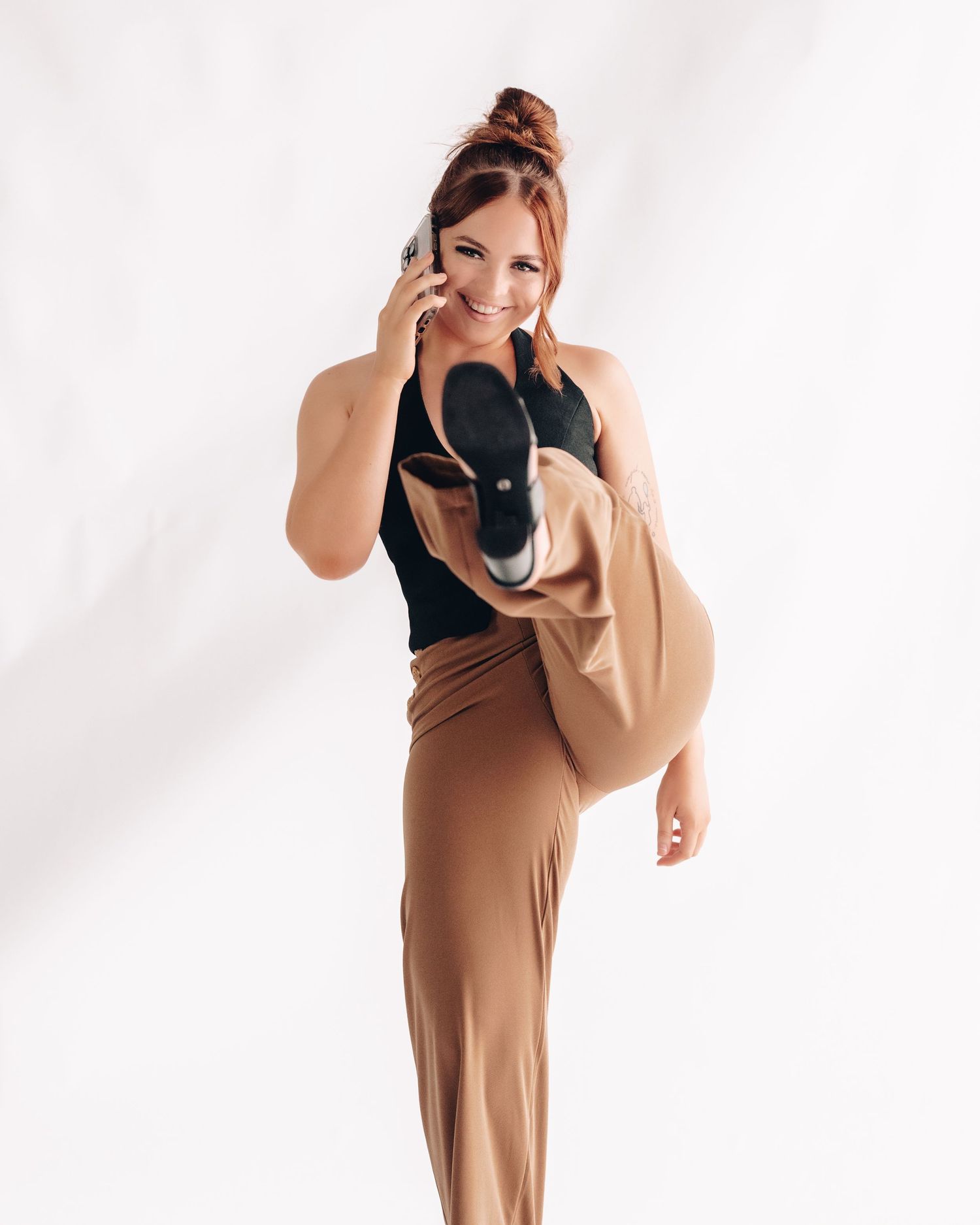Smiling woman in a black top and tan trousers talks on her phone while lifting one leg high in a playful pose.