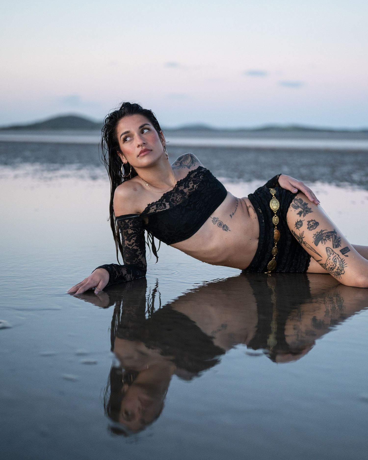 Tattooed woman in black lace outfit lies on a reflective shoreline at dusk, her mirrored image in the shallow water.