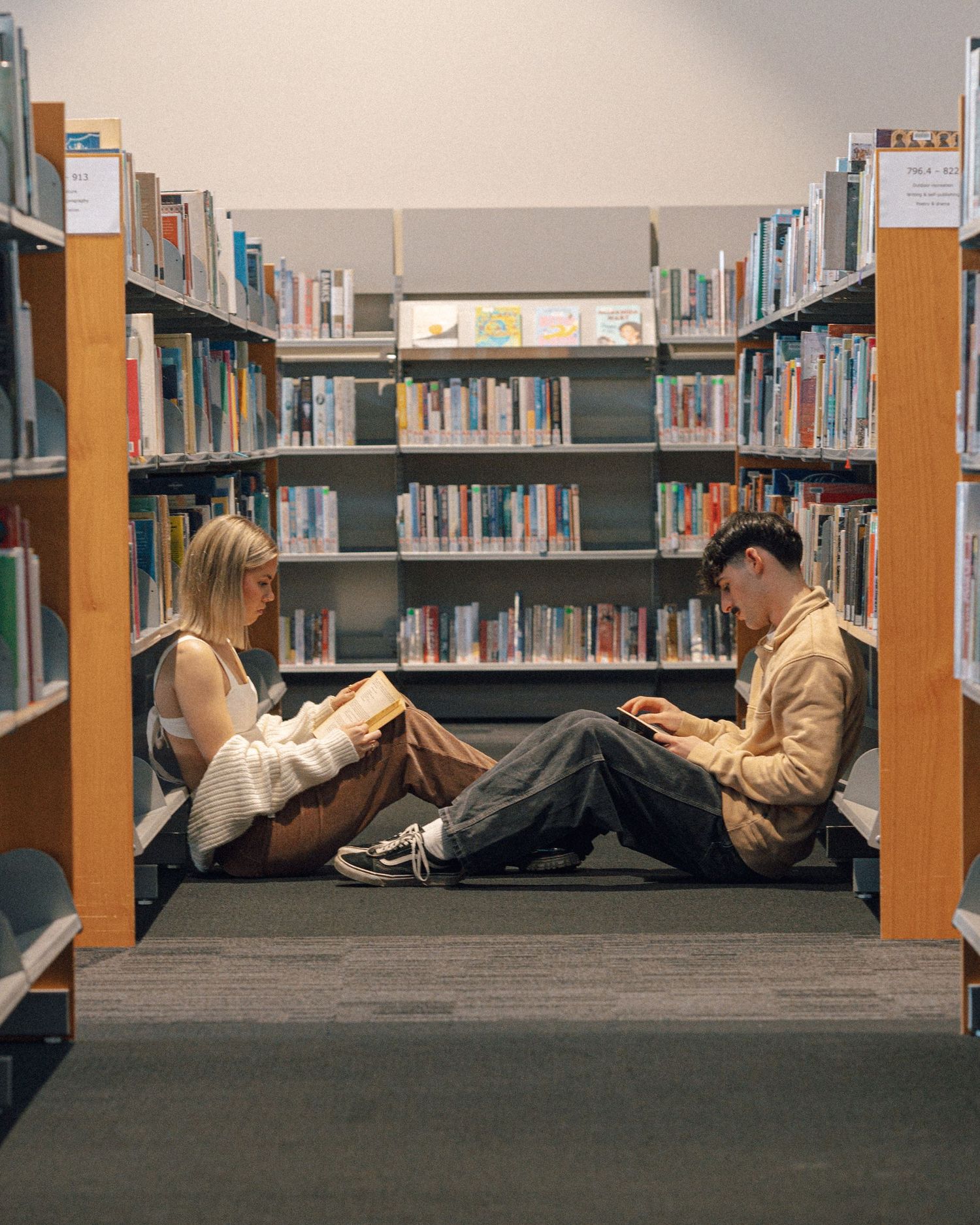 Couple sit on the carpeted floor at opposite ends of a library aisle, quietly reading among tall bookshelves.