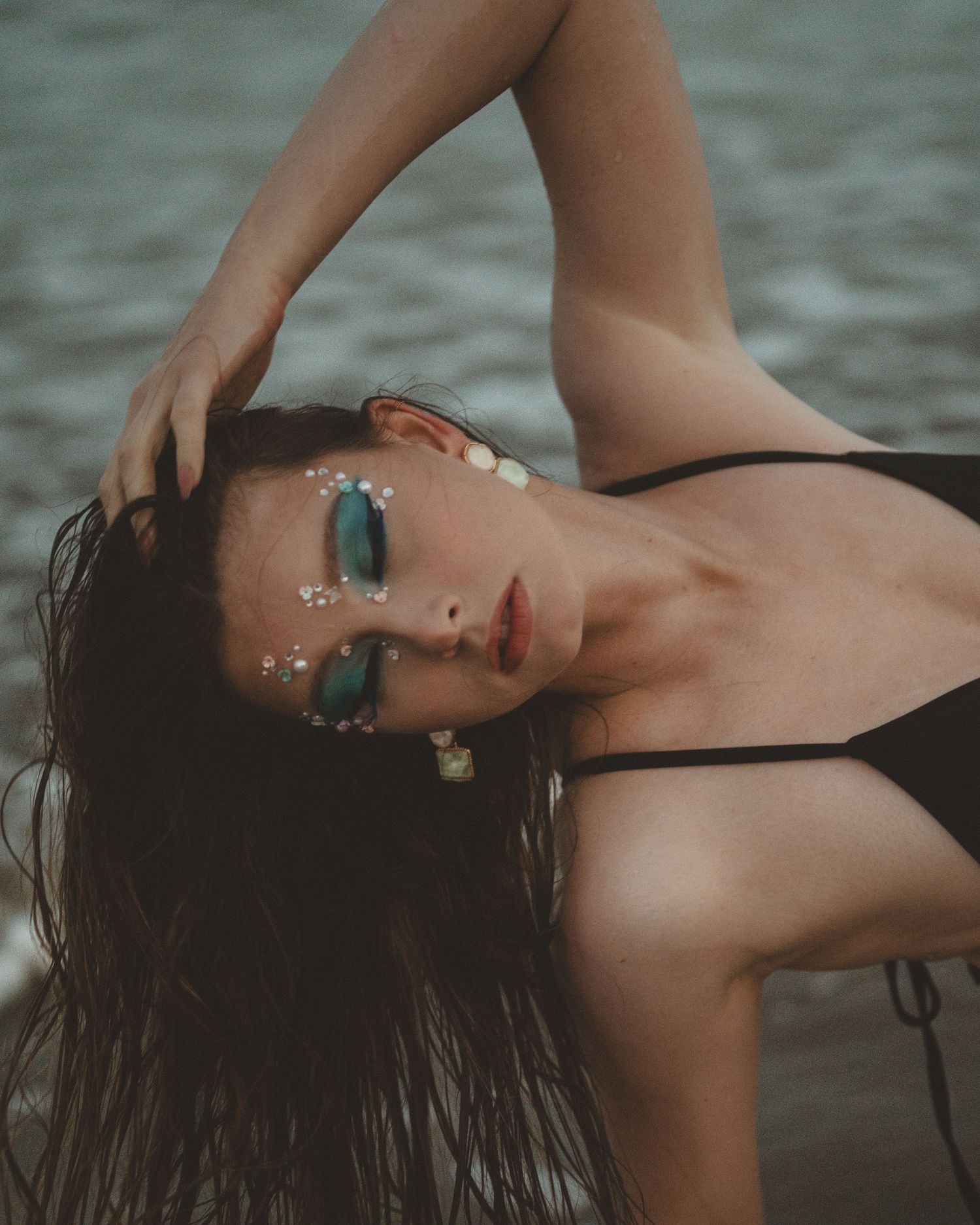 Woman with wet hair leans sideways on the shore, wearing a black swimsuit, teal eyeshadow and rhinestone face gems.