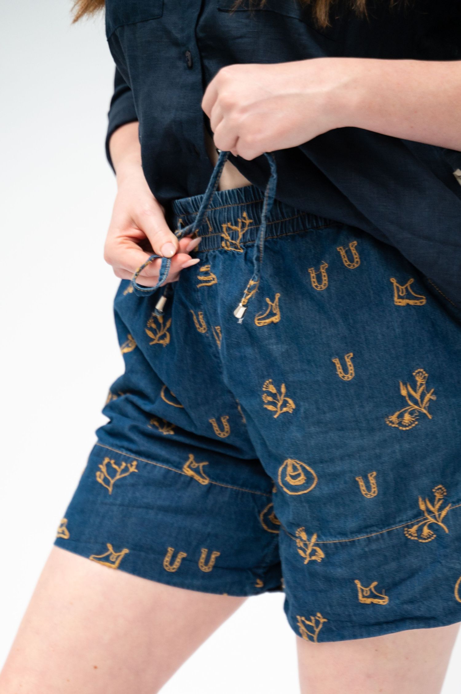 Person in a black button-up shirt tying the drawstring of blue denim shorts embroidered with tan horseshoes, boots and cacti.