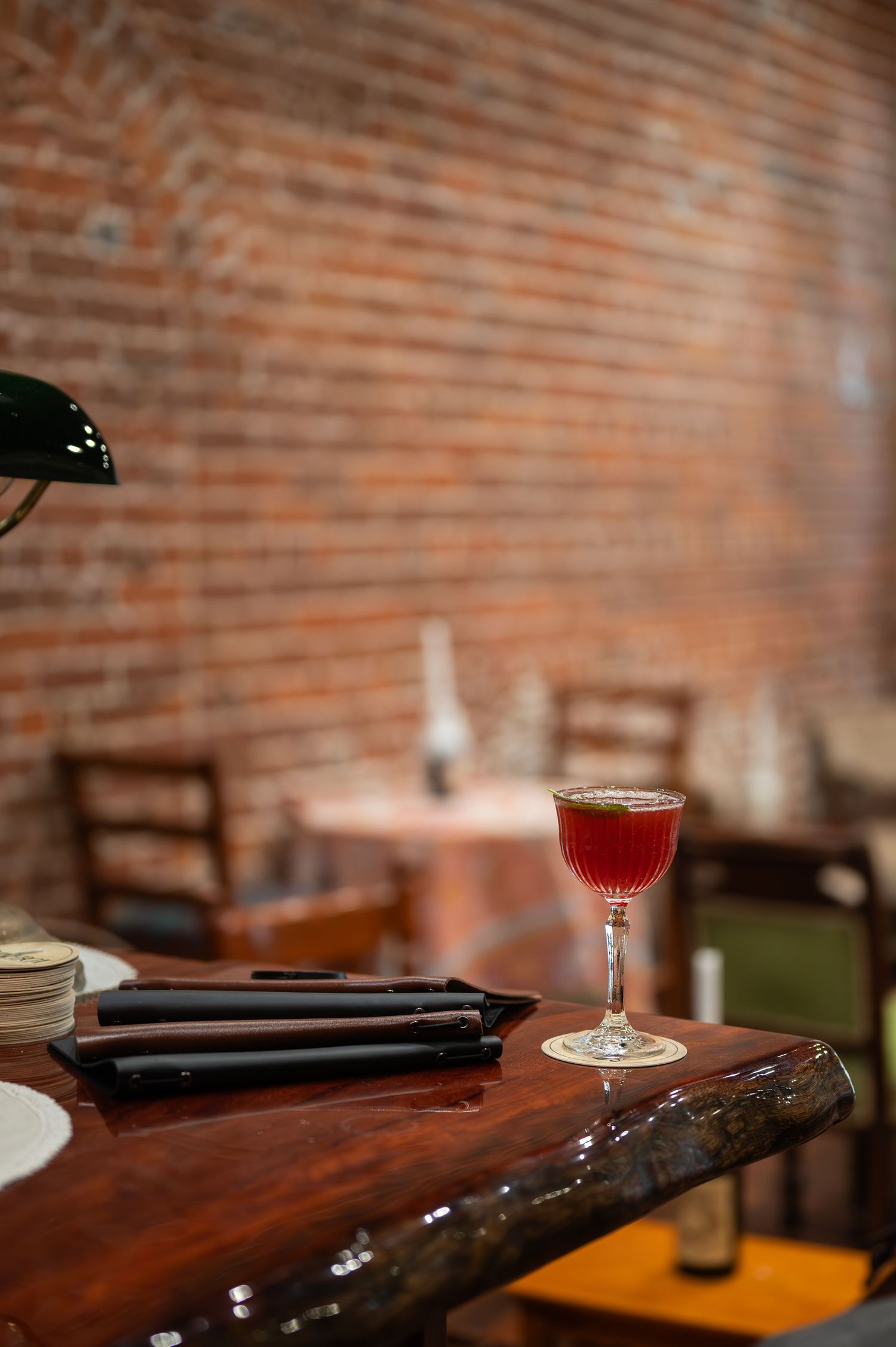 A ruby red cocktail in a coupe glass rests on a live-edge wooden bar top with a brick wall and dining chairs behind.