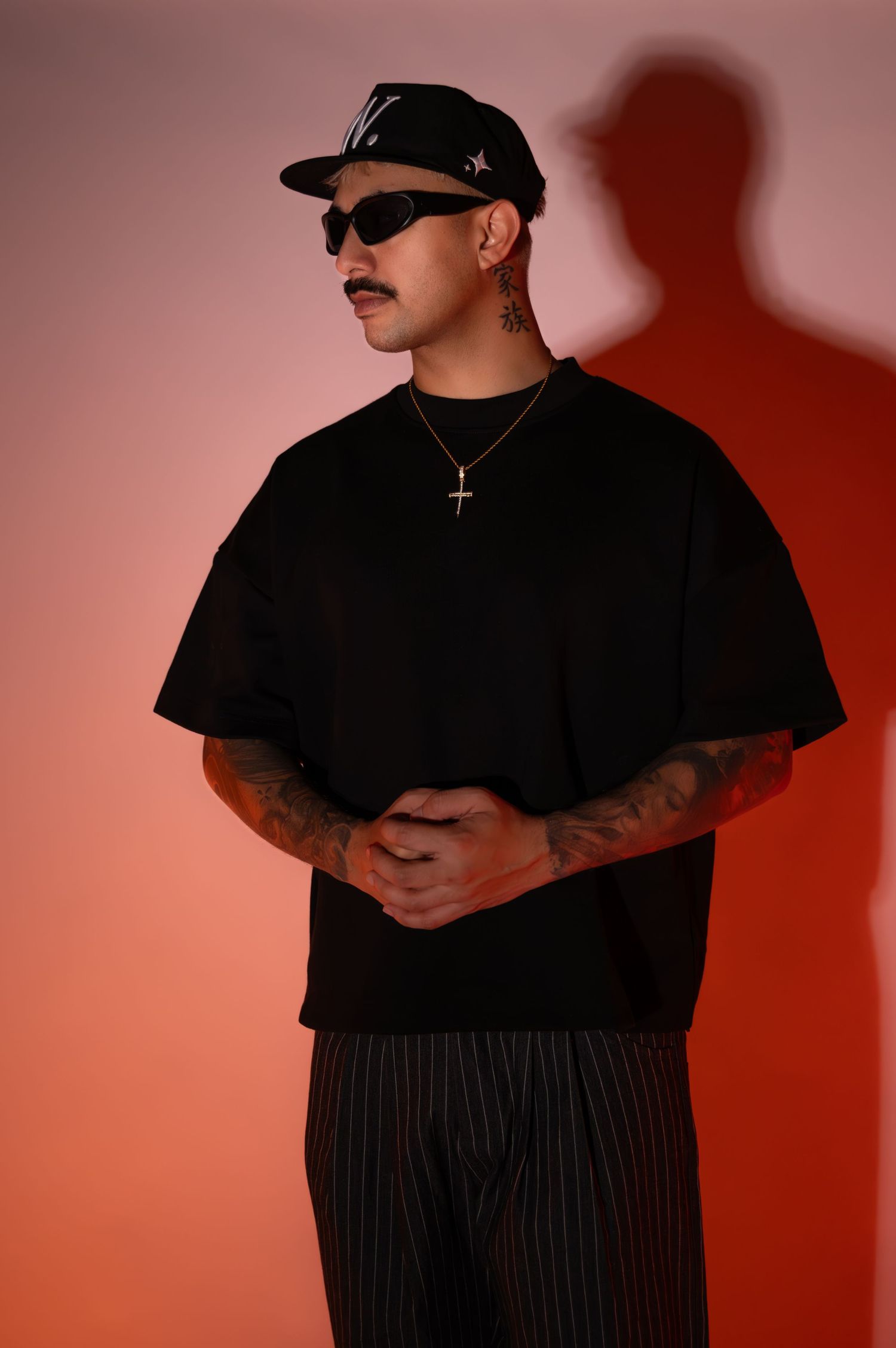 Tattooed man in a black cap, sunglasses, cross necklace and black oversized tee stands with clasped hands against a warm orange backdrop.