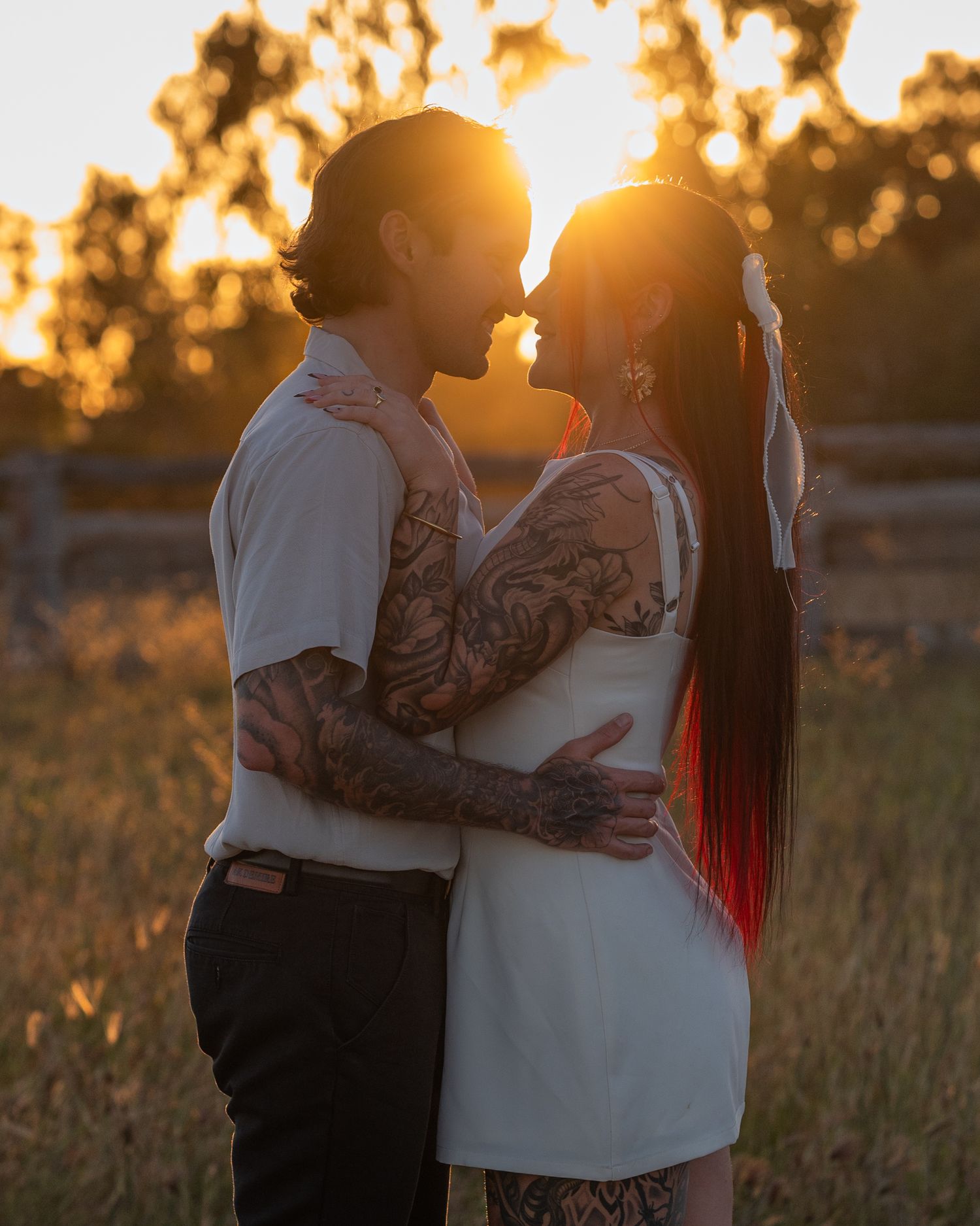 Tattooed couple embraces at golden hour in a grassy field, their faces nearly touching as the sunset glows behind them.