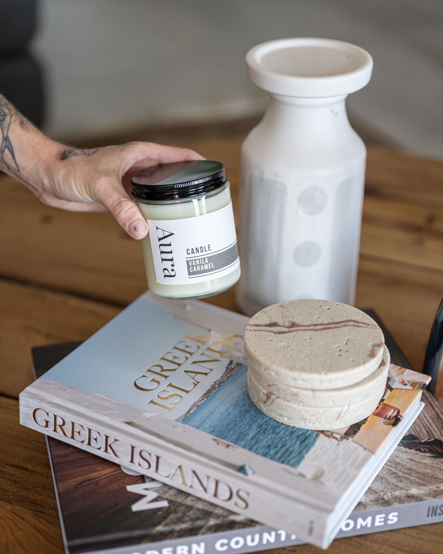 Hand holding an Aura scented candle above coffee table books on Greek Islands, with a white vase and stone coasters.