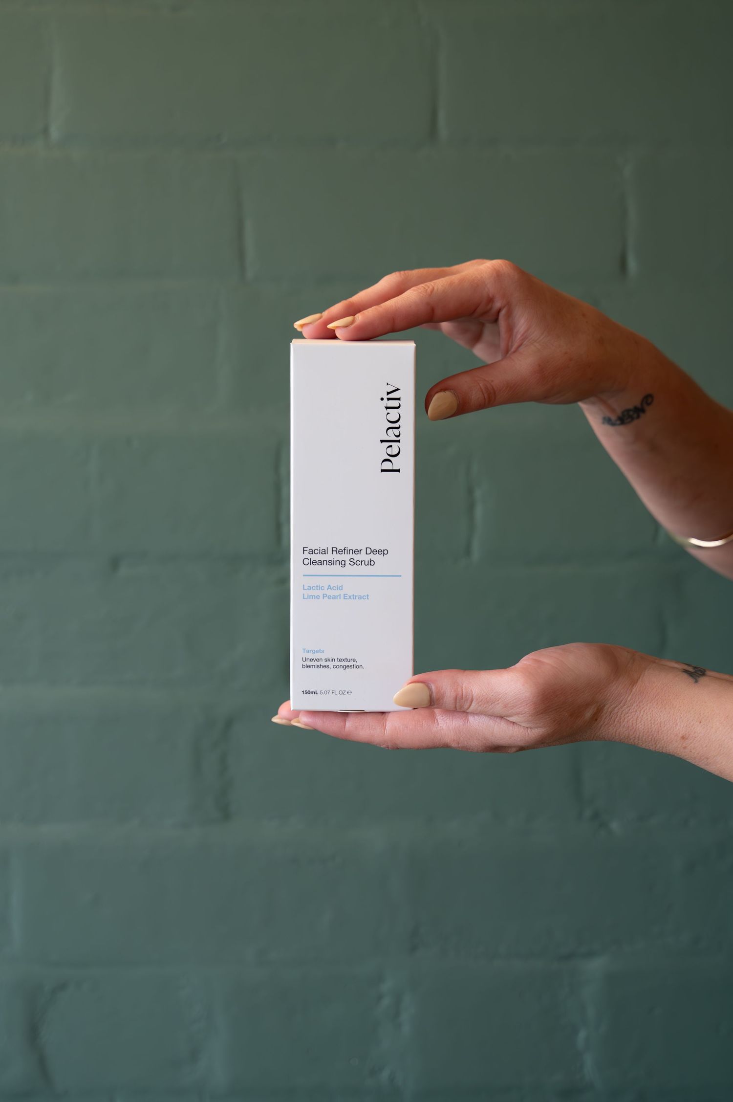 Two hands hold a white Pelactiv Facial Refiner Deep Cleansing Scrub box upright in front of a green painted brick wall.