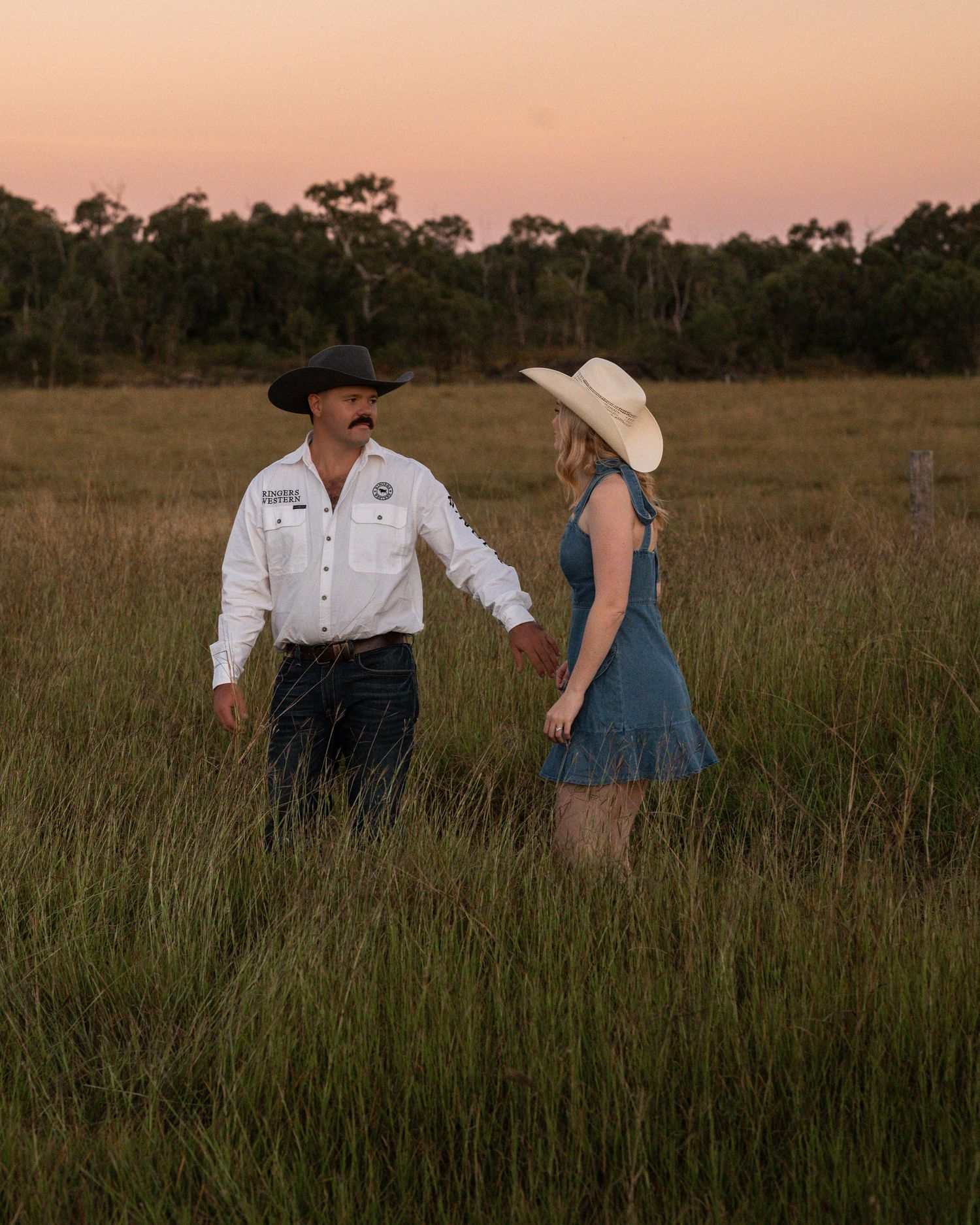 A cowboy and a woman in a straw hat hold hands in a grassy paddock under a soft pink sunset sky.