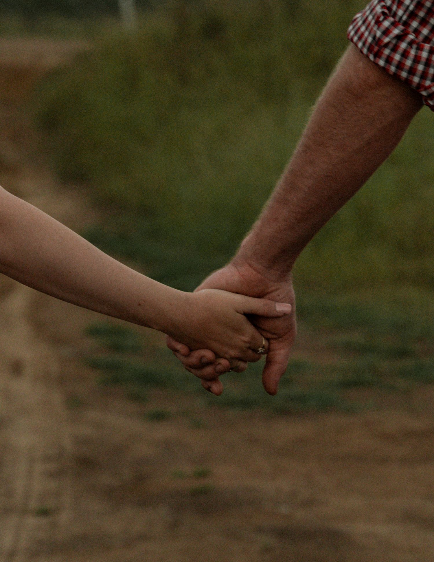 Close-up of a couple holding hands on a dirt country path, with the man in a red plaid shirt sleeve and a ring visible.