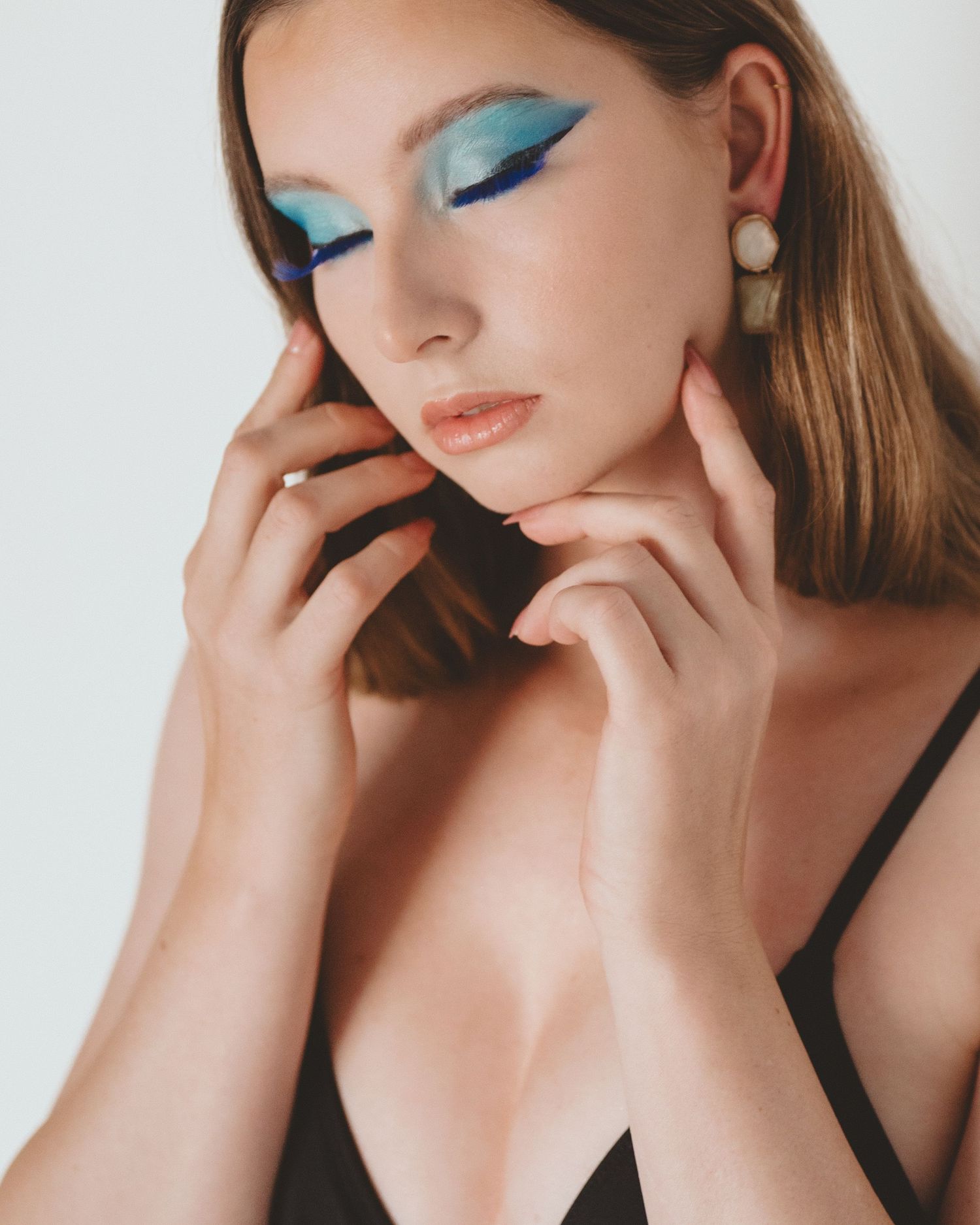 Close-up beauty portrait of a woman with blue winged eyeshadow, pearl drop earrings and hands framing her face in soft natural light.