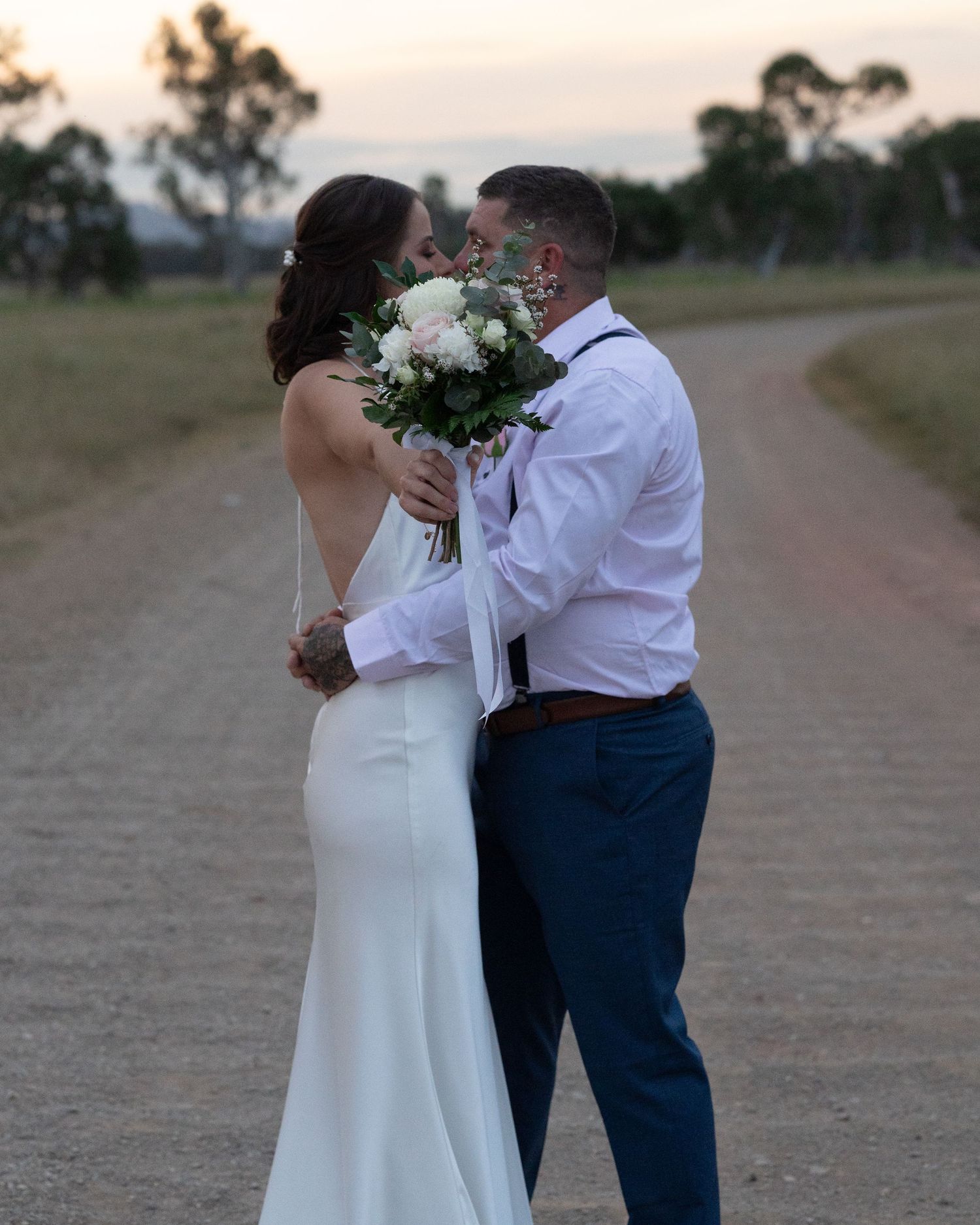 Bride and groom kiss behind a white bouquet on a gravel country road with trees and fields in the background.