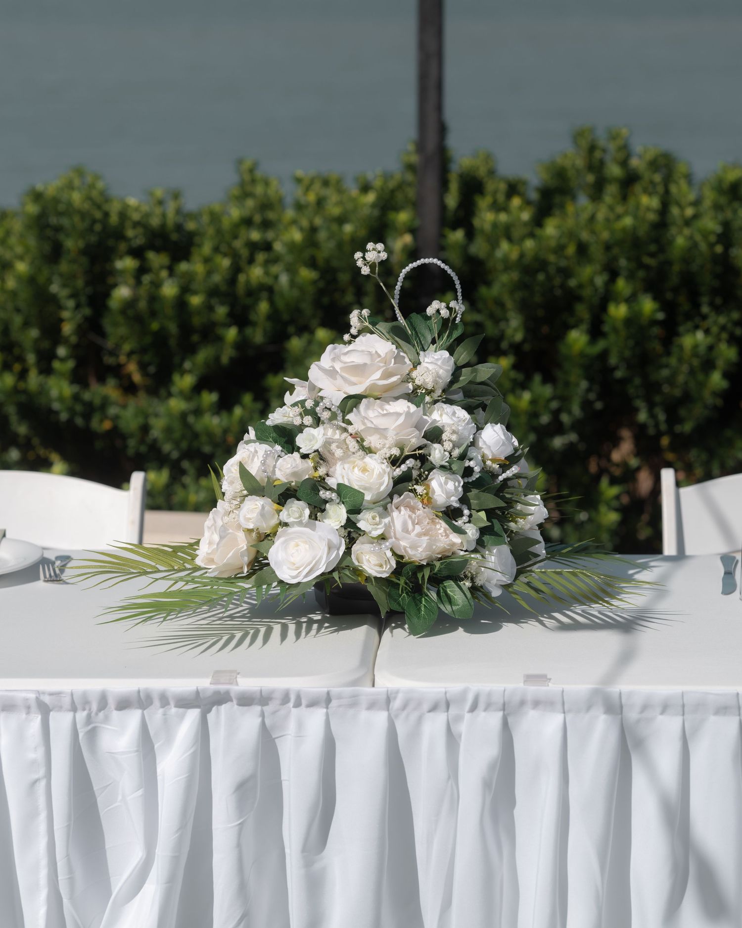 White rose and greenery arrangement with palm fronds sits on a skirted head table at a wedding reception.