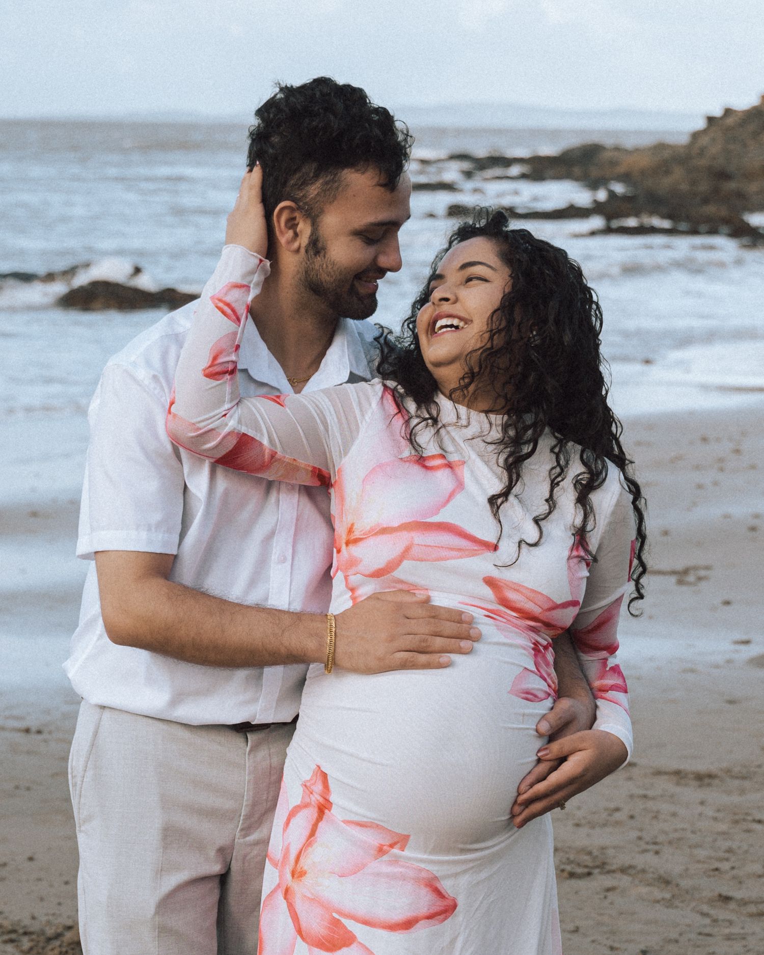 Laughing couple embrace on a rocky beach during a maternity shoot, hands resting on her pregnant belly.
