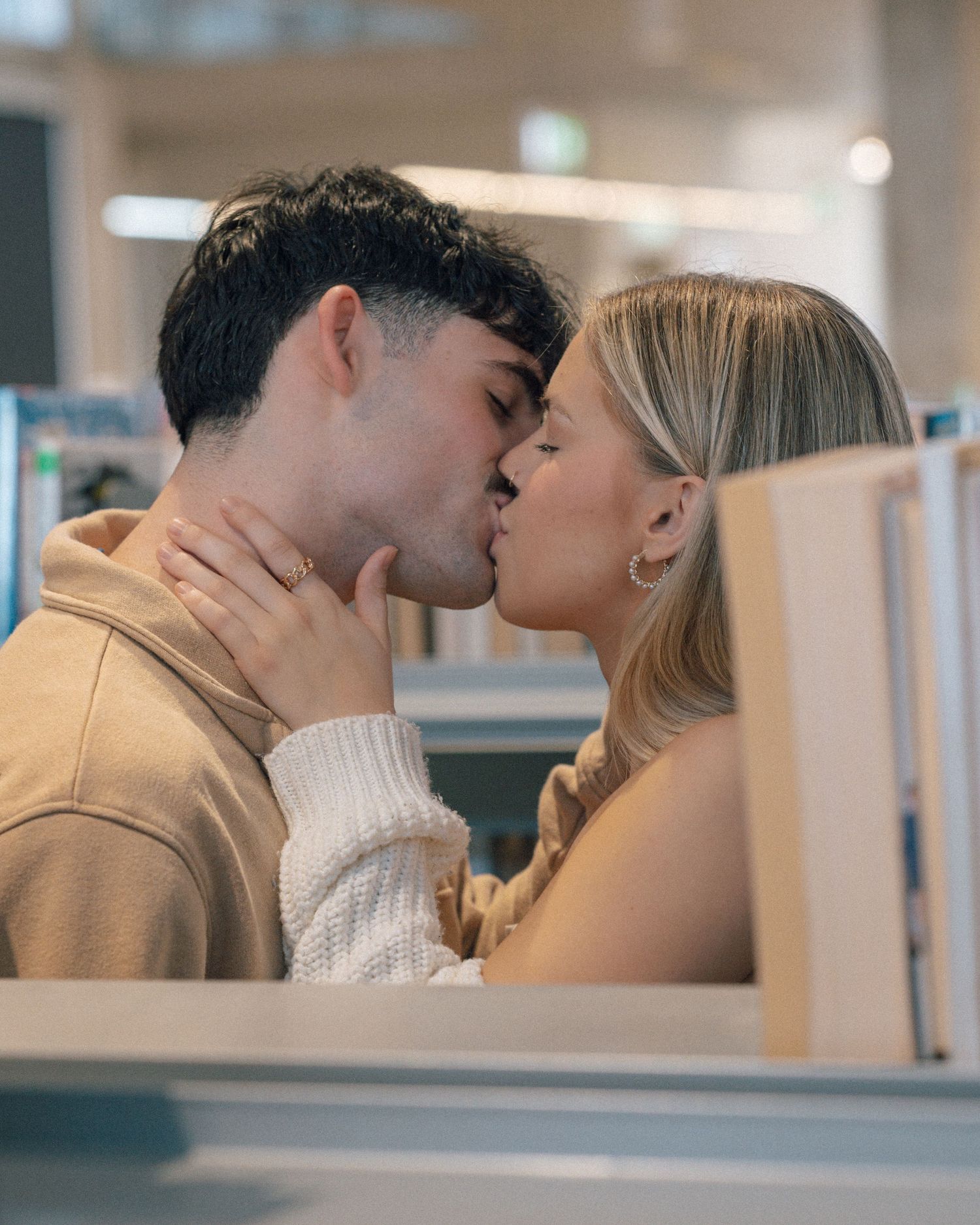 A couple shares a romantic kiss between library shelves, the woman wearing a white knit sweater.