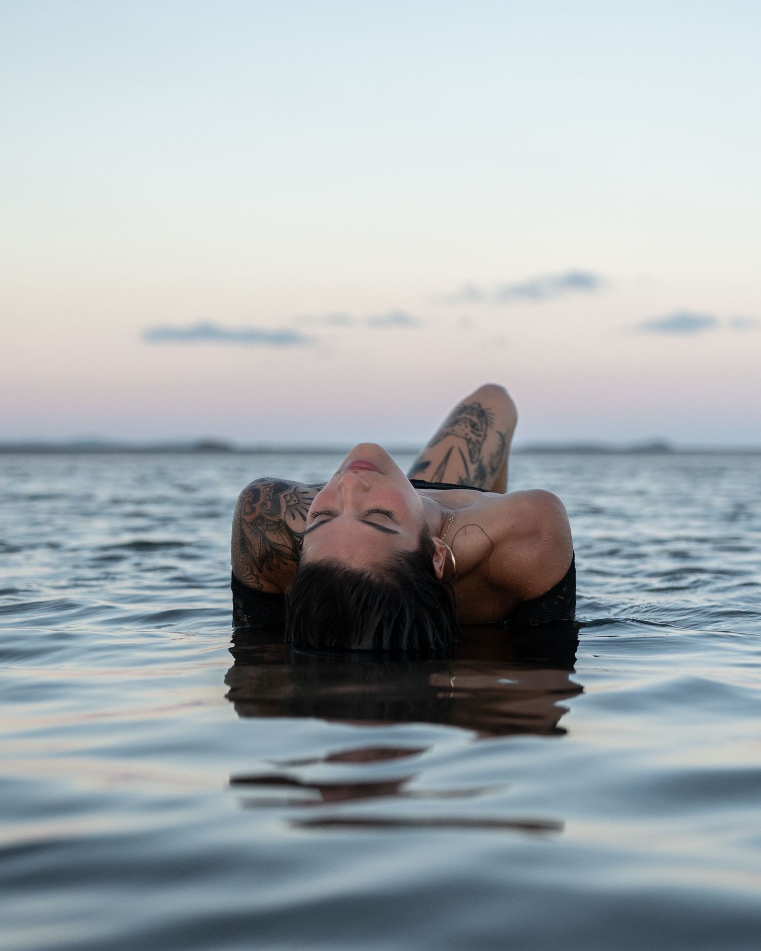 Tattooed woman floating on her back in calm shallow water at dusk with a soft pastel sky and distant shoreline behind her.